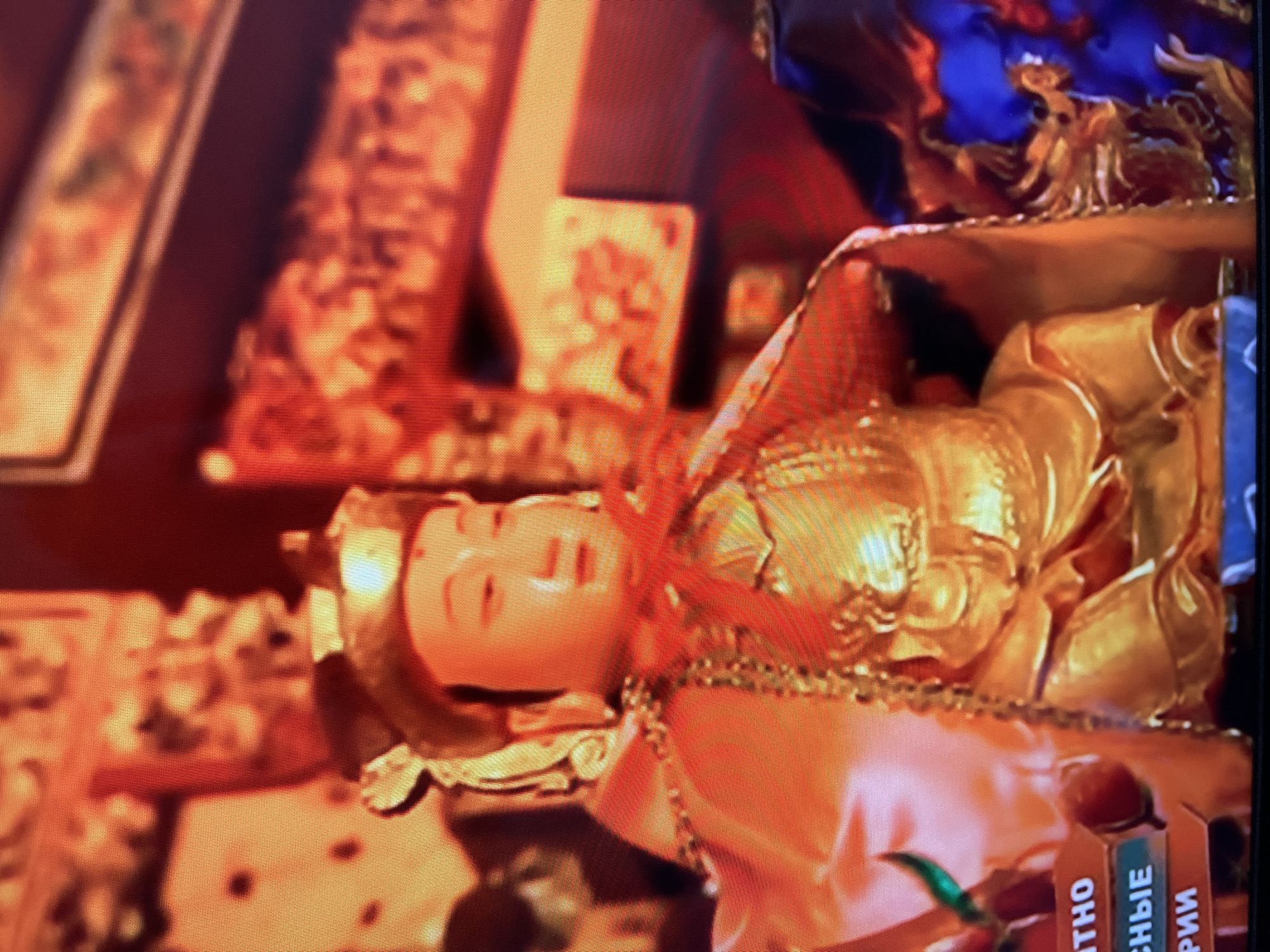 The image displays a richly adorned golden statue, likely depicting a Buddha or Bodhisattva, with a serene expression and closed eyes. The figure wears an elaborate golden headdress and garments, suggesting a regal or divine status. The background features intricate decorative patterns in warm golden and reddish-brown tones, possibly part of a temple interior, shrine, or ornate screen. A vibrant blue element, possibly fabric or a structural component, is visible in the upper right corner, above the statue's head.
The image itself is a photograph taken of a screen, identifiable by the visible pixel grid. It is oriented sideways, showing the statue horizontally, although it would likely be viewed upright in its original context. No activity or event is taking place; it's a static depiction of the statue.
In the bottom right corner, fragments of Cyrillic text are visible on orange-colored graphic overlays. These partial words include "ТНО", "СНЫЕ", and "РИИ", indicating Russian language content. This text likely originates from a broadcast overlay, such as a channel logo, subtitle, or category label, consistent with the provided location context of Moscow, Russia. The overall impression is one of cultural and religious significance.
