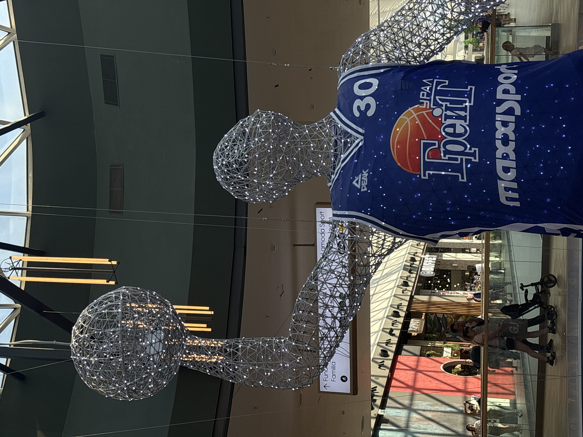 The image depicts a large, decorative sculpture of a basketball player made from wire mesh and illuminated with small lights. The sculpture is wearing a basketball jersey with the number "30" and the logos of "URAL," "Урал," "ТРЕЙЛ" in Cyrillic, "Trail," and "Maxisport." The jersey is predominantly blue and features a basketball graphic.
The sculpture appears to be located inside a shopping mall or similar large indoor space. The architecture includes high ceilings with visible beams and lighting fixtures. Reflections on glass barriers or floors suggest the presence of retail stores and pedestrian traffic below.
Given the Cyrillic lettering and logos, the location is identified as the city of Perm, Russia, as indicated by the logo for the basketball team "Ural Great Perm" (Урал-Грейт Пермь).