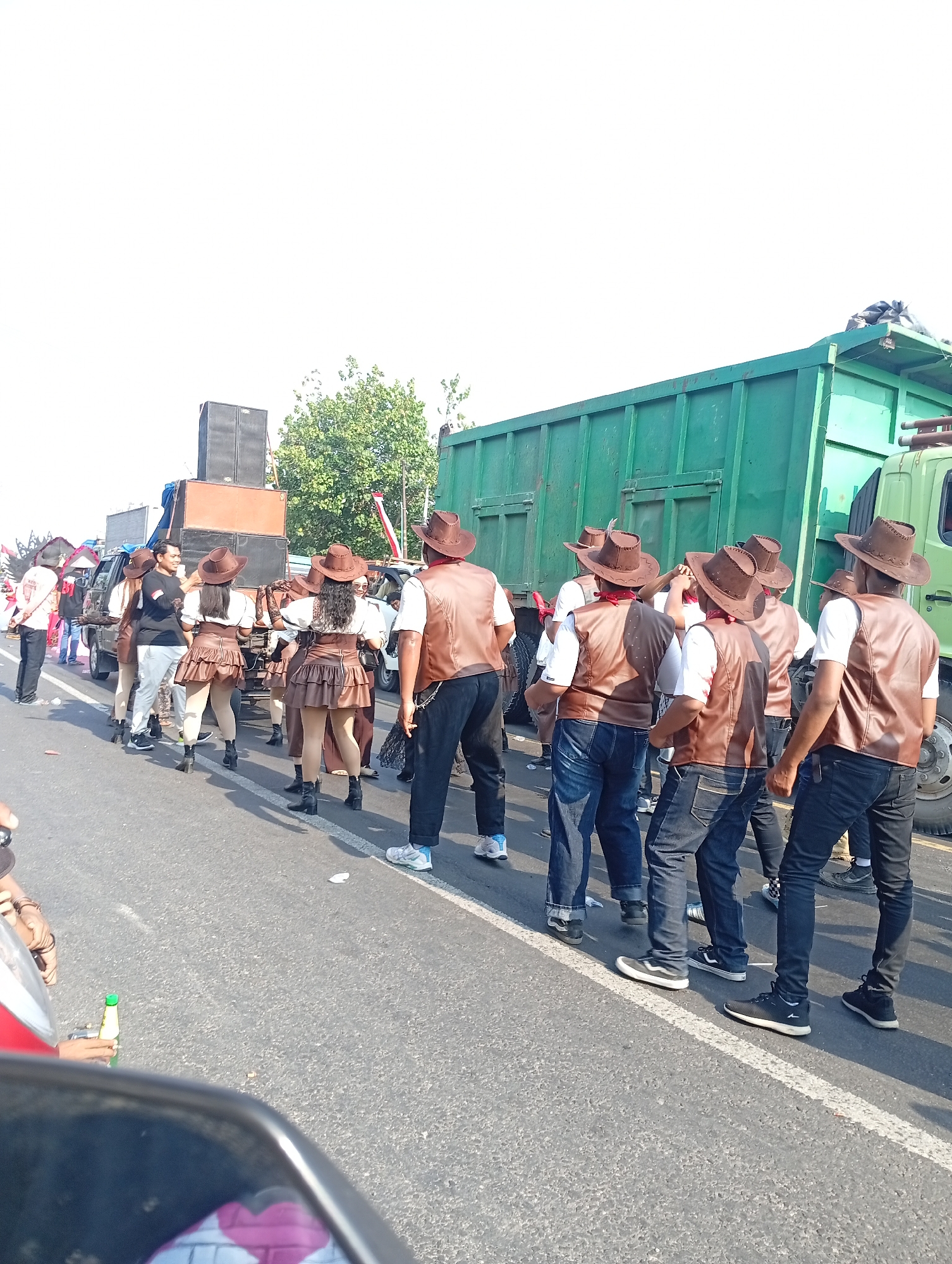 The image depicts a street scene in Sarang, Indonesia, likely during a parade or performance. A group of individuals, predominantly men, are seen from behind, all wearing brown cowboy hats and matching brown leather vests over white shirts. Several women are also present, dressed in brown cowboy hats, white blouses, and tiered brown skirts. They appear to be part of the same procession, walking along a paved road.
In the background, a large green truck with a dump bed is parked on the right side of the road. To the left, a smaller vehicle, possibly a van, is visible, loaded with large speakers stacked high. Behind the speakers, more people are gathered. The environment appears to be an urban or semi-urban setting, with a bright, overcast sky. The road is marked with a white line. The overall impression is of a lively event taking place outdoors.