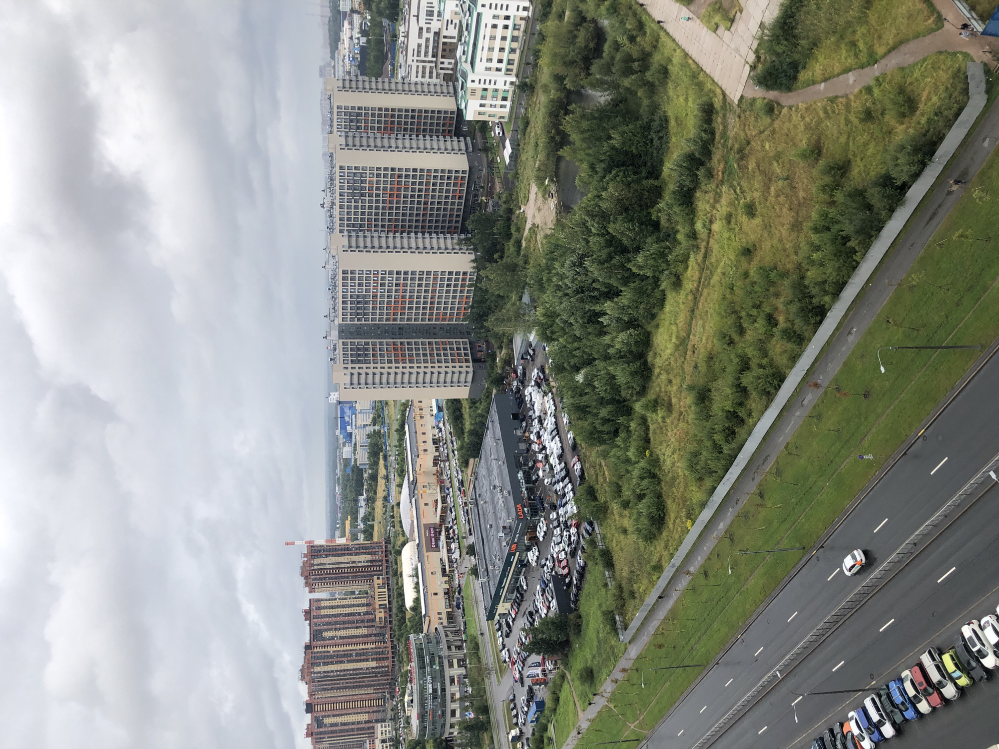 An aerial shot captures a modern urban landscape under a cloudy sky. In the foreground, a multi-lane road runs diagonally from the bottom left, with parked cars lining the left side. Several cars are visible driving on the road.
Beyond the road, a large, dark-roofed building with "LADA" signage is prominent, surrounded by a vast parking lot filled with vehicles. To the left of this LADA dealership is a curved, multi-story building with a glass facade, labeled "TOKYO-CITY". Further back, a sprawling complex of beige and orange buildings with numerous windows stretches across the frame, appearing to be residential high-rises. Adjacent to these are more modern apartment buildings with white and green facades.
A patch of dense green trees and foliage occupies the middle ground, bordering a small body of water. The background reveals more buildings and industrial structures, with a tall, striped red and white smokestack visible in the distance. The overall scene depicts a contemporary urban environment with significant commercial and residential development.