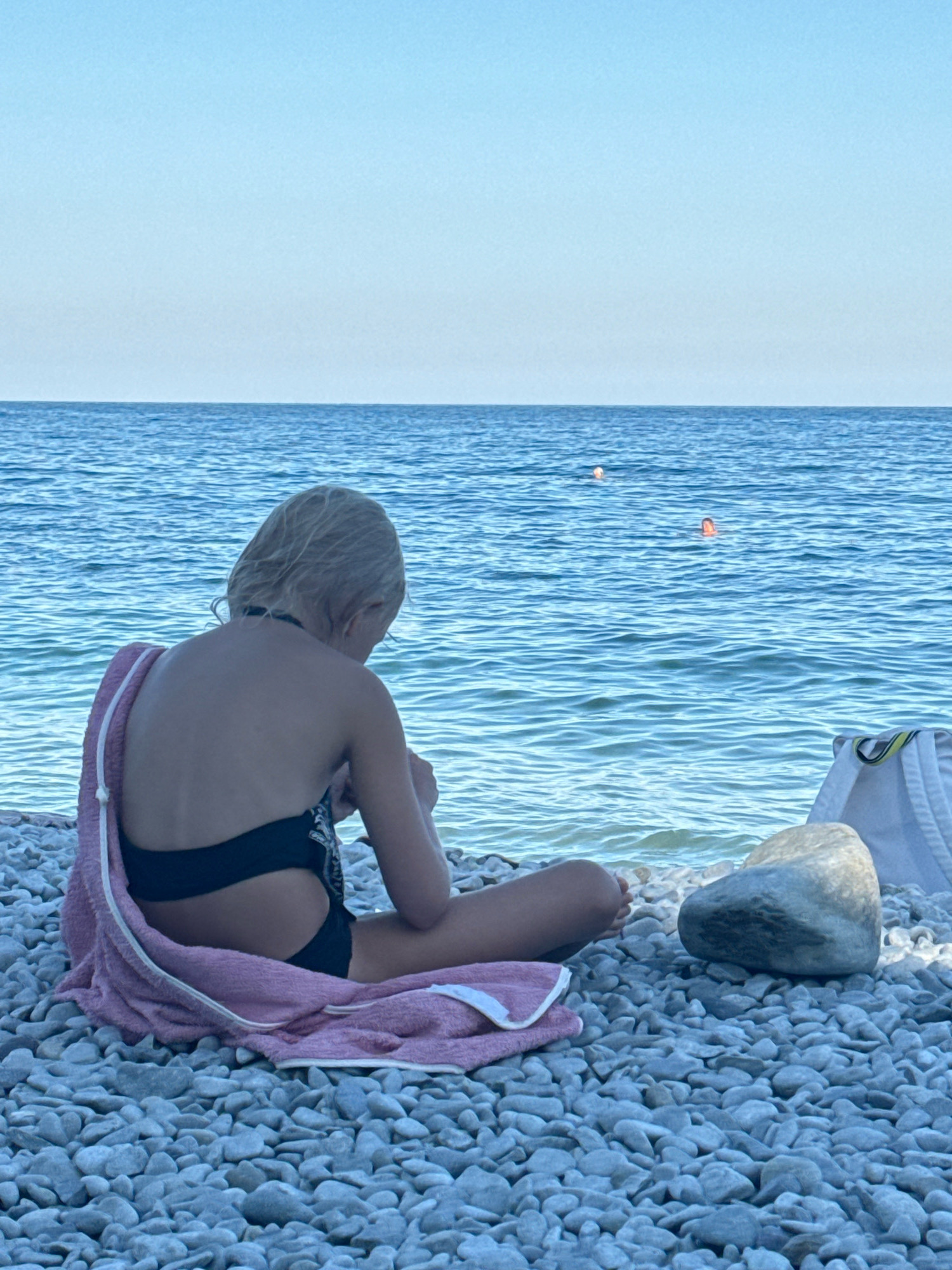 A young girl with blonde hair, viewed from behind, sits on a pebble beach, facing the sea. She wears a black bikini and is wrapped in a pink towel draped over her shoulders. The girl is looking down and appears to be holding something in her hands. The sea is a calm, bright blue, with gentle ripples. In the distance, two people are visible swimming in the water. The sky is a clear, pale blue, suggesting a sunny day. The beach is covered in smooth, light-colored pebbles. A white beach bag and a large, smooth grey rock are near the girl. The overall impression is one of a peaceful summer day at the seaside. The time of day appears to be afternoon, given the bright sky and clear visibility. No text is visible in the image. The scene is set in Partenit, Ukraine.