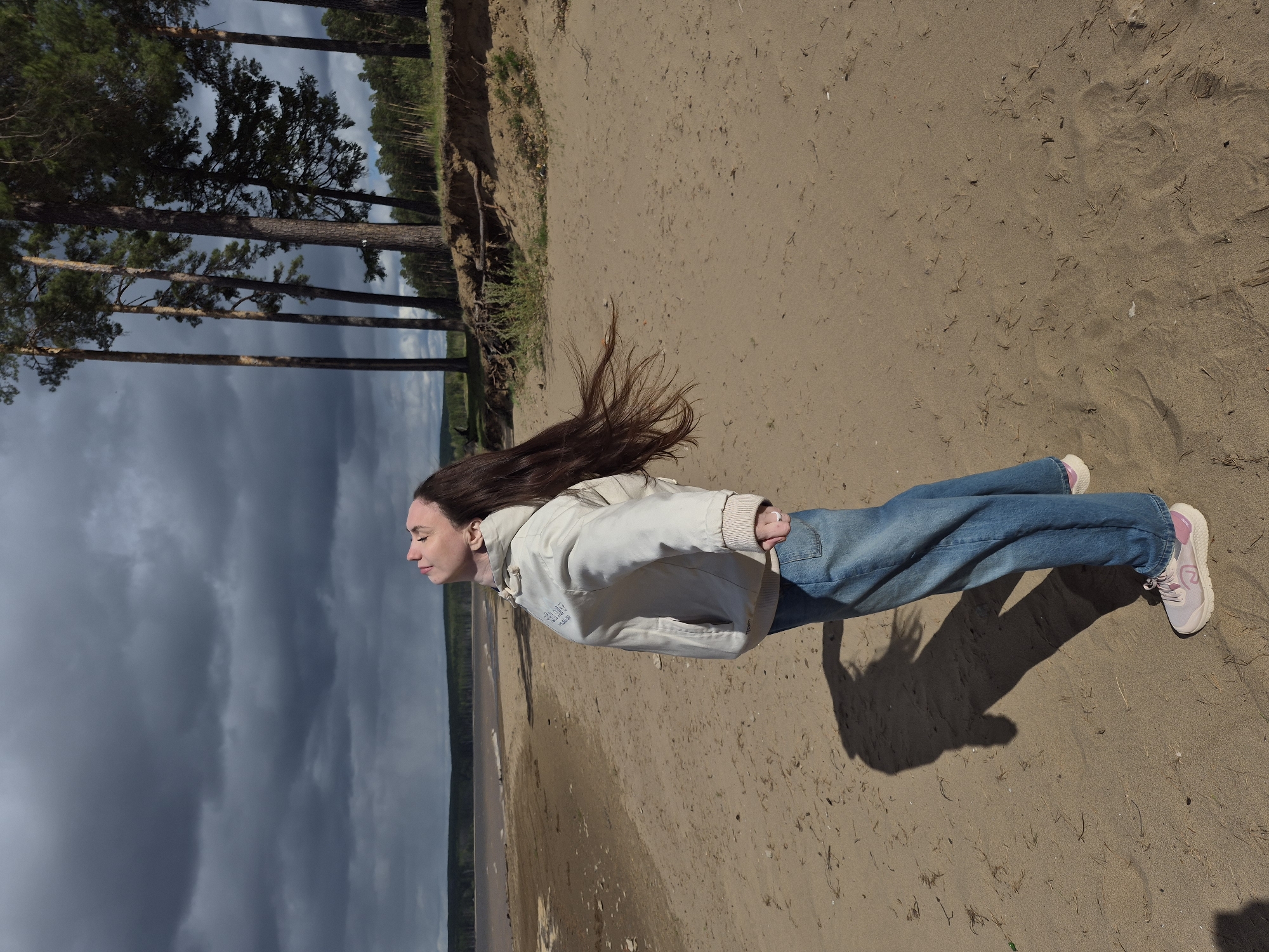 A woman is lying face down in the sand on the shore of the Kama River in the city of Chaikovsky, Russia. She is wearing blue jeans, a white jacket, and pink sneakers. The woman's long brown hair is splayed out on the sand. In the background, there is a line of trees and a cloudy sky.