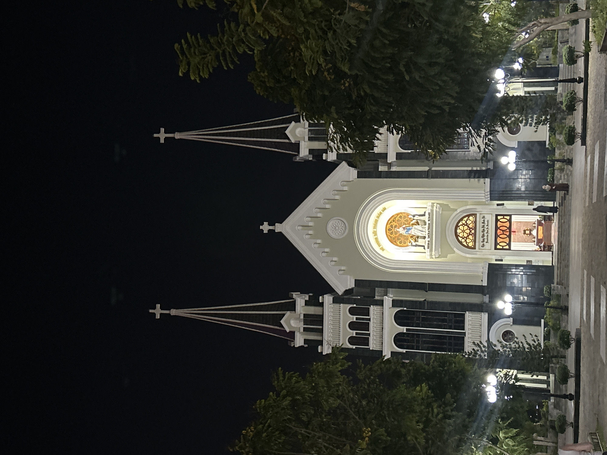 The image shows the Nha Trang Cathedral in Bắc Nha Trang, Vietnam, at night. The cathedral, a light-colored stone structure with dark accents, features two bell towers topped with crosses, flanking a central gable. The building is illuminated, highlighting its arched windows, decorative stone work, and a large circular stained-glass window above the entrance. The cathedral's doors are open, revealing the interior. In front of the cathedral, lampposts are spaced along a walkway, creating pools of light. Trees are visible, framing the cathedral and partly obscuring some of its features. Two people are visible walking near the building entrance.