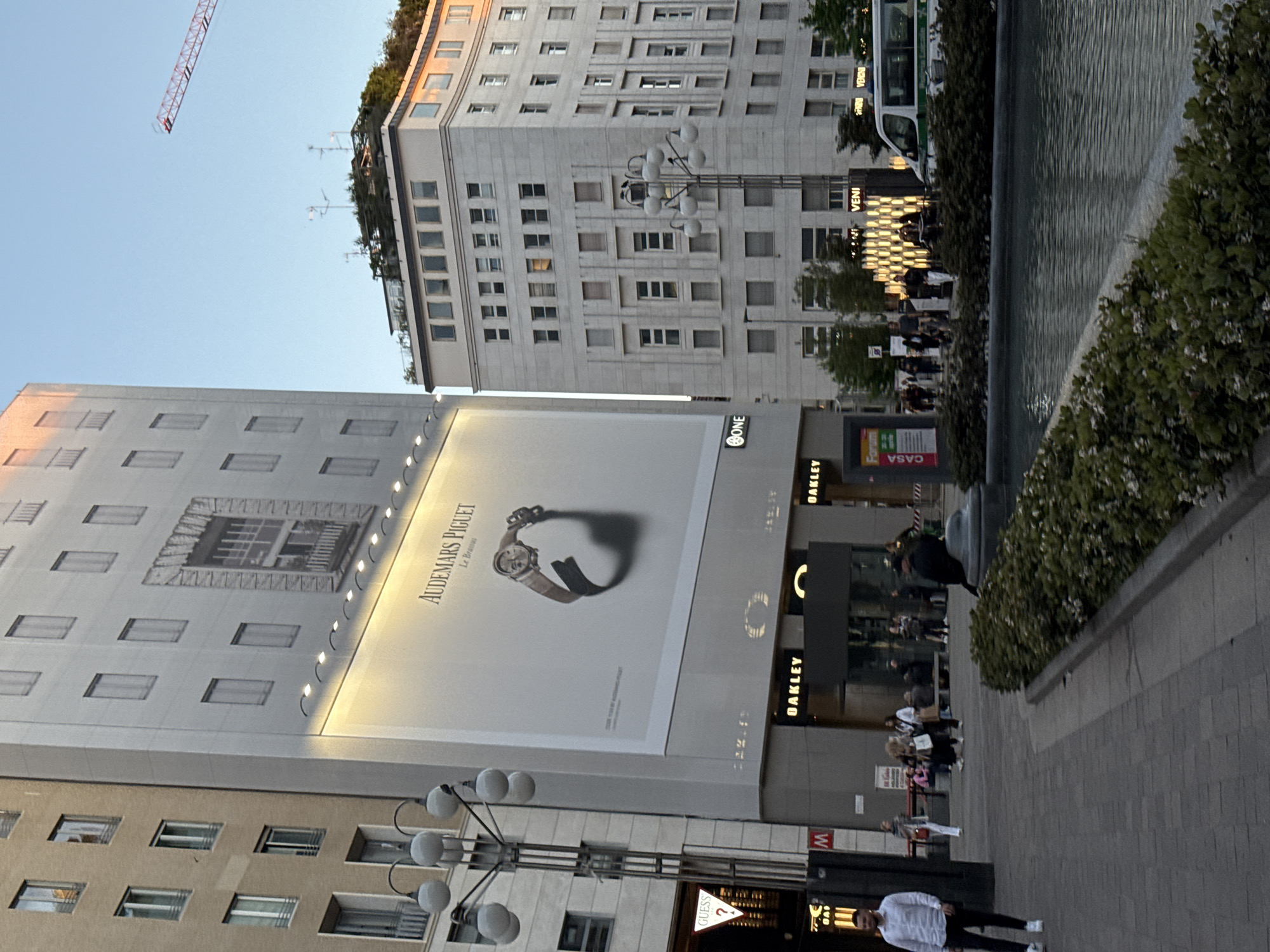 This media file captures an urban street scene in Milan, Italy, depicting a bustling area with prominent commercial buildings and public spaces. The image appears to be taken in the late afternoon or early evening, indicated by the clear, light blue sky and the soft, diffused lighting.
On the left, a tall, light-colored building features numerous small rectangular windows and is largely covered by a massive, brightly illuminated billboard. The billboard advertises "AUDEMARS PIGUET Le Brassus" and displays a large image of a luxury watch with a brown leather strap. Below the billboard, storefronts with signs for "ONE" and "OAKLEY" are visible. Further down, at street level, a "GUESS ?" sign and a "M" (likely for a metro station) sign can be seen.
In the middle-right of the frame stands a multi-story building constructed of light grey stone, characterized by its numerous windows and a somewhat ornate facade. The rooftop of this building is adorned with greenery, suggesting a rooftop garden or terraced landscaping, and also features several antennas. A multi-headed street lamp with spherical globes is positioned in front of this building.
To the far right, a narrow water feature, possibly a fountain or canal, runs parallel to the street. Bushes and small trees line its edge, separating it from the paved pedestrian walkway. In the background, beyond the water, parts of a white and green bus are visible, with text like "VENI" discernible on its side. Several people are scattered across the scene; some are walking along the sidewalk, while others appear to be congregating near the storefronts. A man in a white shirt and dark pants walks prominently in the foreground. A red construction crane is visible in the upper left corner against the clear sky, hinting at ongoing development in the area. Other visible text includes "Forum C CASA" on a small sign near the sidewalk.