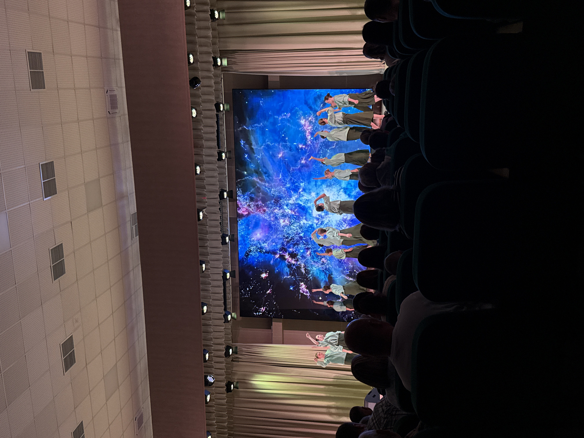 The image shows a scene inside a theater in Veliky Novgorod, Russia. A digital screen displays an image of dancers against a blue and purple galaxy background. There are rows of seated audience members in the foreground. Along the left wall of the theatre are rows of lights. The lighting is dim.
