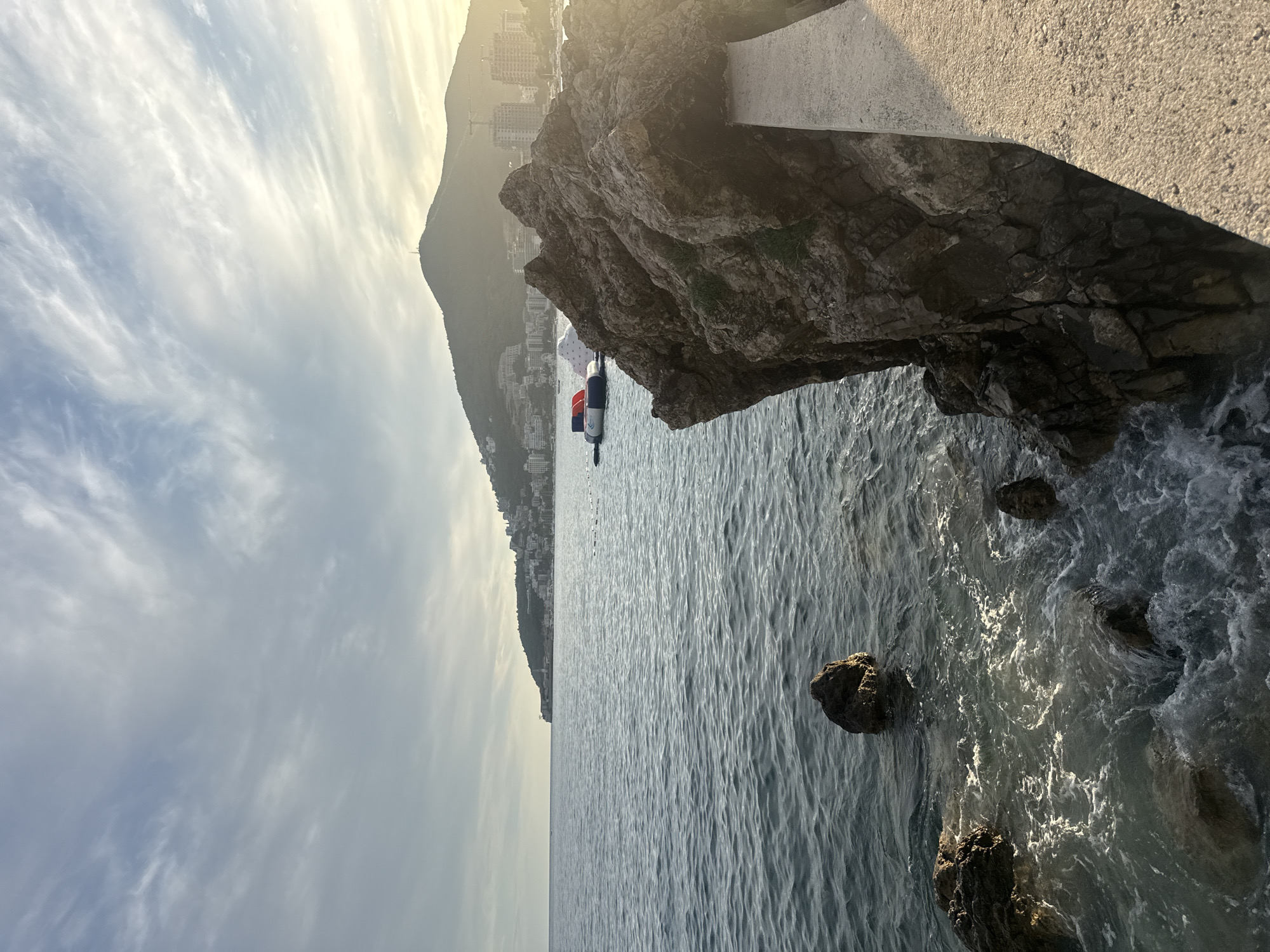 The image captures a scenic view of the Adriatic Sea with a rocky shore in the foreground and the city of Budva, Montenegro, in the background. The sun is setting, casting a warm glow over the landscape.
In the foreground, large, rugged rocks jut out into the sea, with waves gently crashing against them. A concrete walkway or pier runs along the right side of the frame, leading towards the rocks. The water is a deep blue, with white foam where the waves meet the rocks.
The middle ground of the image features a calm expanse of the sea, with a few buoys and what appear to be inflatable water toys floating on the surface. In the distance, the city of Budva is visible, nestled along the coastline and climbing up a verdant hill. Numerous buildings, including hotels and apartments, are spread across the hillside, and a prominent antenna sits atop the hill. Construction cranes are also visible, indicating ongoing development in the city.
The background is dominated by a large, silhouetted hill, with the sky above displaying a mix of wispy clouds and clear blue. The setting sun, partially obscured by the hill, creates a soft, diffused light that illuminates the clouds and casts a warm hue on the scene. The overall impression is one of tranquility and natural beauty, juxtaposed with the urban landscape of Budva.