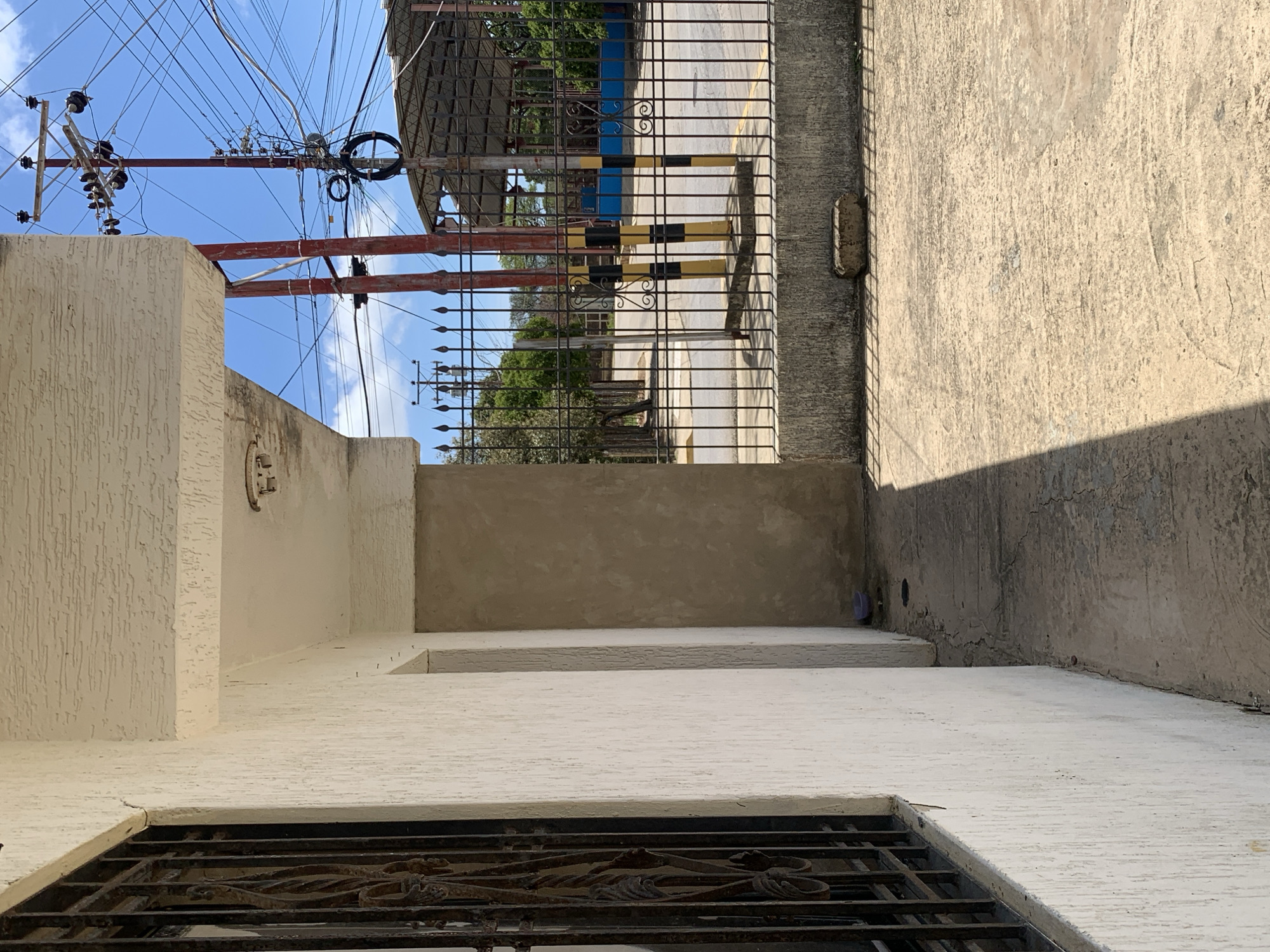 The image depicts a street view in Santa Ana, Venezuela. A light-colored, textured wall of a building, with a black metal-barred window on the left, extends towards the foreground and right. Adjacent to this wall is a covered walkway or entryway with a white ceiling and a beige column. Beyond the building, a black metal fence with pointed finials encloses a space. Through the fence, a street is visible, with a large number of electrical wires and poles dominating the upper portion of the frame, indicating a densely wired urban environment. Several trees and a covered structure, possibly a market or pavilion, are also visible in the background. The ground in front of the building is paved with concrete, showing cracks and wear, with a clear shadow cast across it by the building. A small purple object, resembling a bowl or container, is on the ground near the base of the building. The sky is blue with scattered clouds. The scene appears to be captured during daylight.