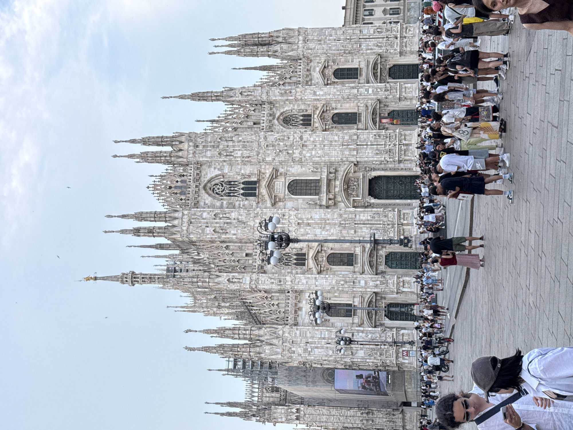 This is a photograph taken from a ground-level perspective, looking up at the Milan Cathedral in Milan, Italy. The cathedral, constructed of white marble, dominates the upper half of the frame with its intricate Gothic architecture, featuring numerous spires, statues, and arched windows. In the foreground, a large crowd of people are gathered, most of whom are casually dressed and appear to be tourists. Some individuals are looking at their phones, while others are talking or observing the cathedral. The ground is paved with large, light-colored stone tiles. The sky is overcast, with some scattered clouds. There are several lampposts visible in front of the cathedral. A large advertisement screen is mounted on the side of the cathedral to the left. The overall scene depicts a busy public square in front of a significant architectural landmark.