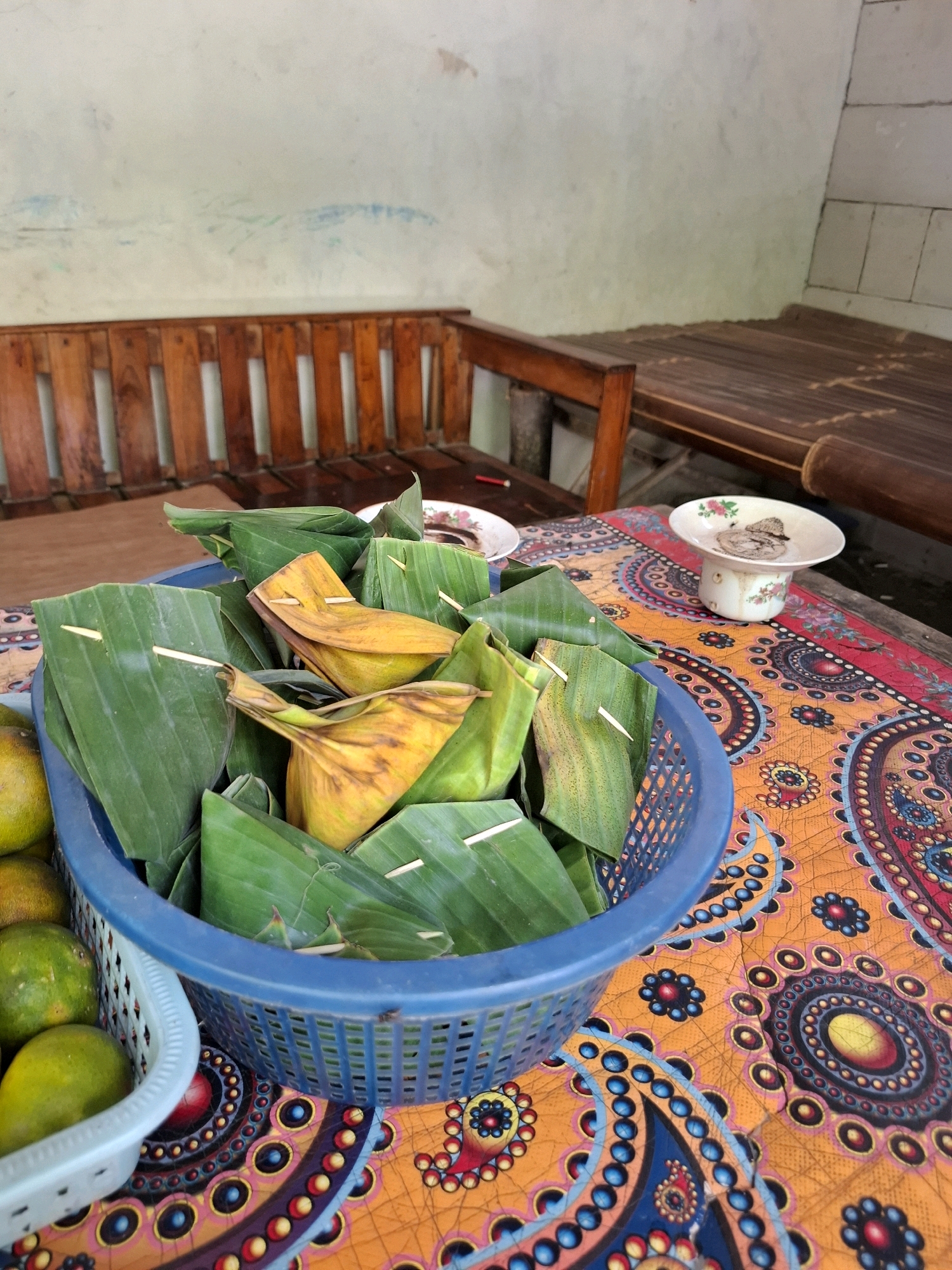 A blue plastic basket filled with food wrapped in banana leaves is positioned on a table with an orange, blue, and yellow patterned cloth. The food items, secured with toothpicks, appear to be small, triangular parcels, some with yellowish hues indicative of being cooked or containing a filling. To the left of the basket, a smaller, lighter blue plastic container holds several green fruits, likely limes or small oranges. In the background, a wooden bench and a table with a bamboo surface are visible. The wall behind the furniture is a pale, textured surface, possibly plaster. The scene suggests an outdoor or semi-outdoor setting, possibly a food stall or a home kitchen in Kedungmaron, Indonesia, given the context.