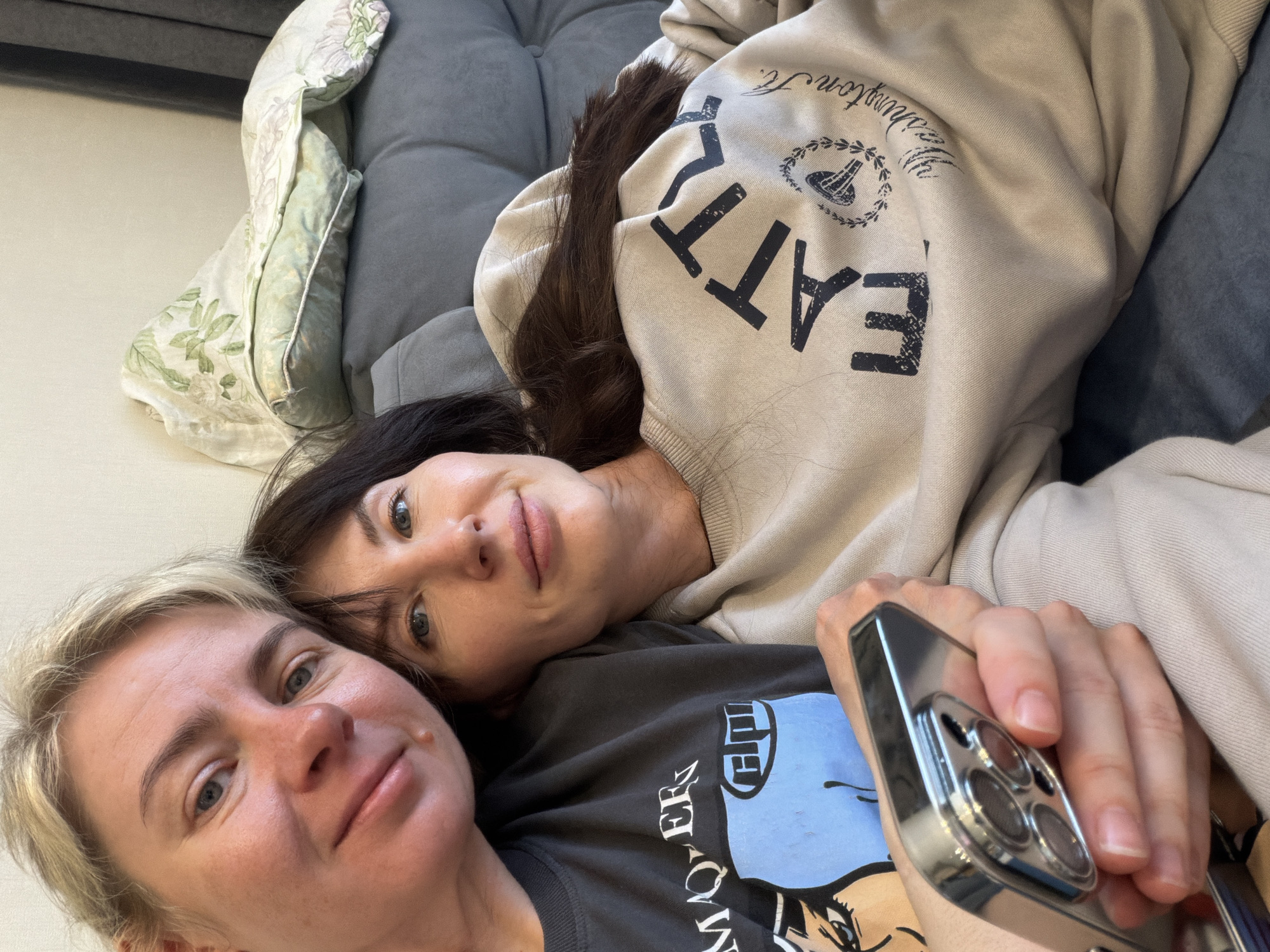 Two Caucasian women are lying on a grey sofa. One woman has blonde hair and is wearing a dark grey t-shirt with a graphic design. She is holding a silver smartphone. The other woman has dark brown hair and is wearing a beige hoodie with the word "EAT" printed on it. A decorative pillow with a floral pattern is visible on the left side of the frame. The location and city of Perm, Russia, cannot be determined from the image.