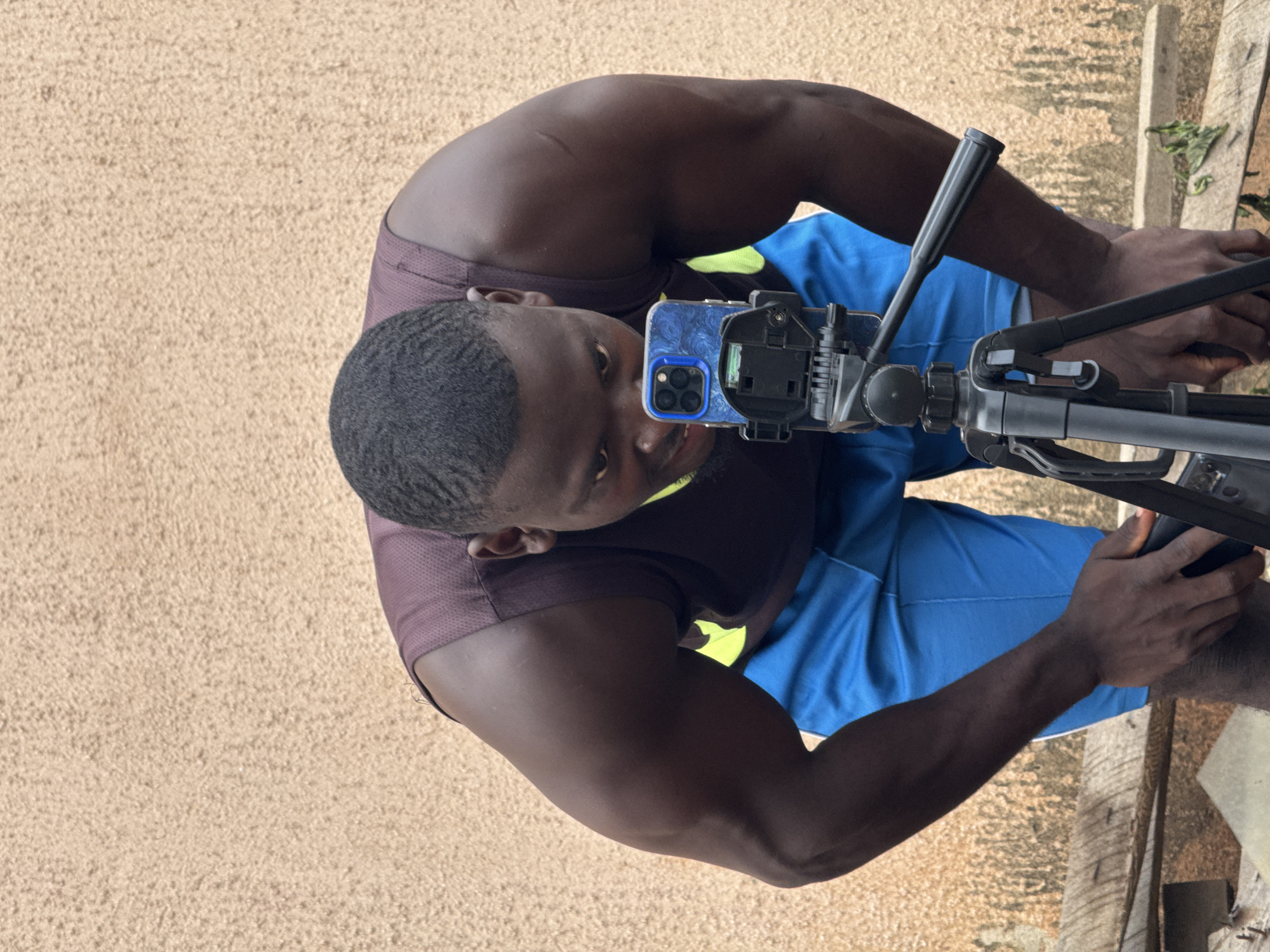 A dark-skinned man is positioned in a low crouch, with his head bent forward as he looks intently at a smartphone mounted on a tripod. The man wears a sleeveless dark purple athletic shirt with neon yellow accents and blue athletic shorts. His muscular arms are visible and he is leaning on the tripod with his right hand. The smartphone has a blue and white patterned case. The background is a textured, light brown wall, possibly stucco, with some wear and tear visible. Wooden planks are present in the foreground and lower portion of the image, suggesting an outdoor or partially constructed area. The scene appears to be in an urban setting, potentially in Akwanga, Nigeria, given the architectural style of the wall and the general environment.