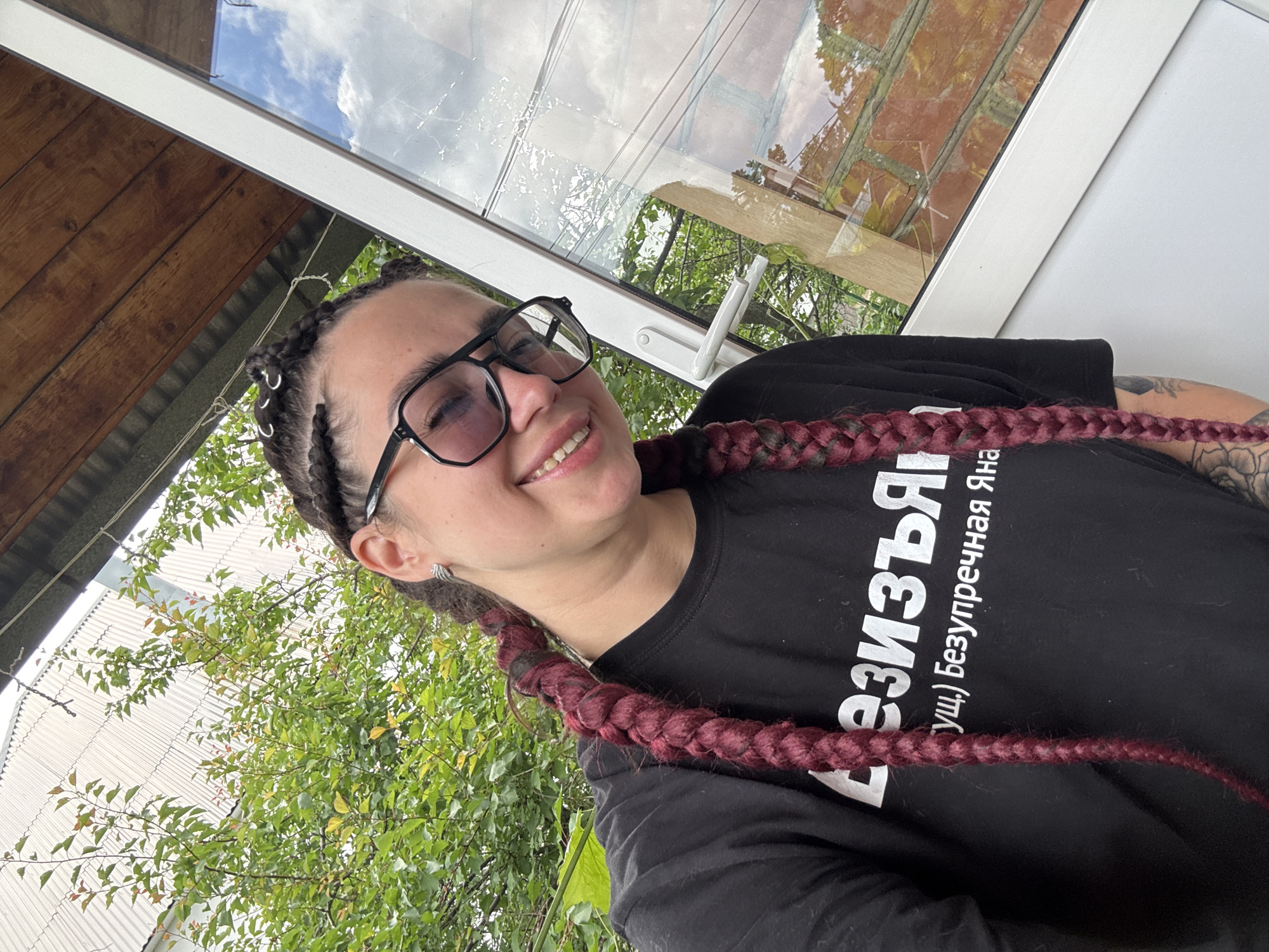 A woman with dark skin, dark braided hair with red highlights, and black-rimmed glasses is taking a selfie outdoors. She is wearing a black t-shirt with white lettering. She is smiling, and her left arm is visible, showing tattoos. She is positioned in front of a glass window, with greenery and a wooden roof visible in the background. The location is described as Unknown, Unknown.