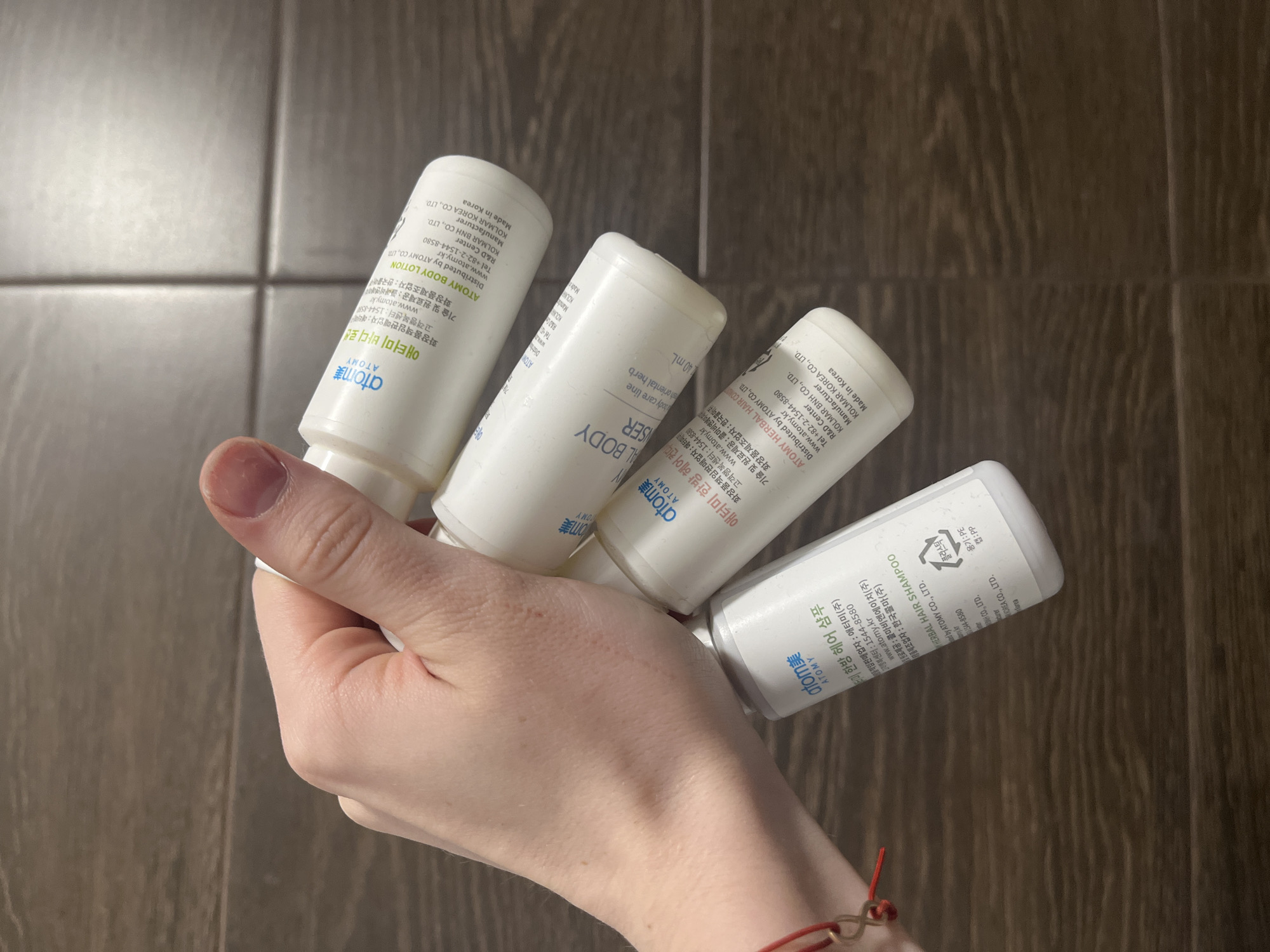 A close-up shows a hand holding four small, white bottles of Atomy brand personal care products. The bottles are labeled, in part, with the brand name "atomy美 ATOMY" and include: "HERBAL HAIR SHAMPOO", "HERBAL HAIR CON", "BODY LOTION", and "SER". The bottles include Korean text, manufacturer information, and recycling symbols. The background is a wood-patterned surface. A small piece of red string is visible on the wrist. There is no indication of the city of Saint Petersburg, Russia, or any location.