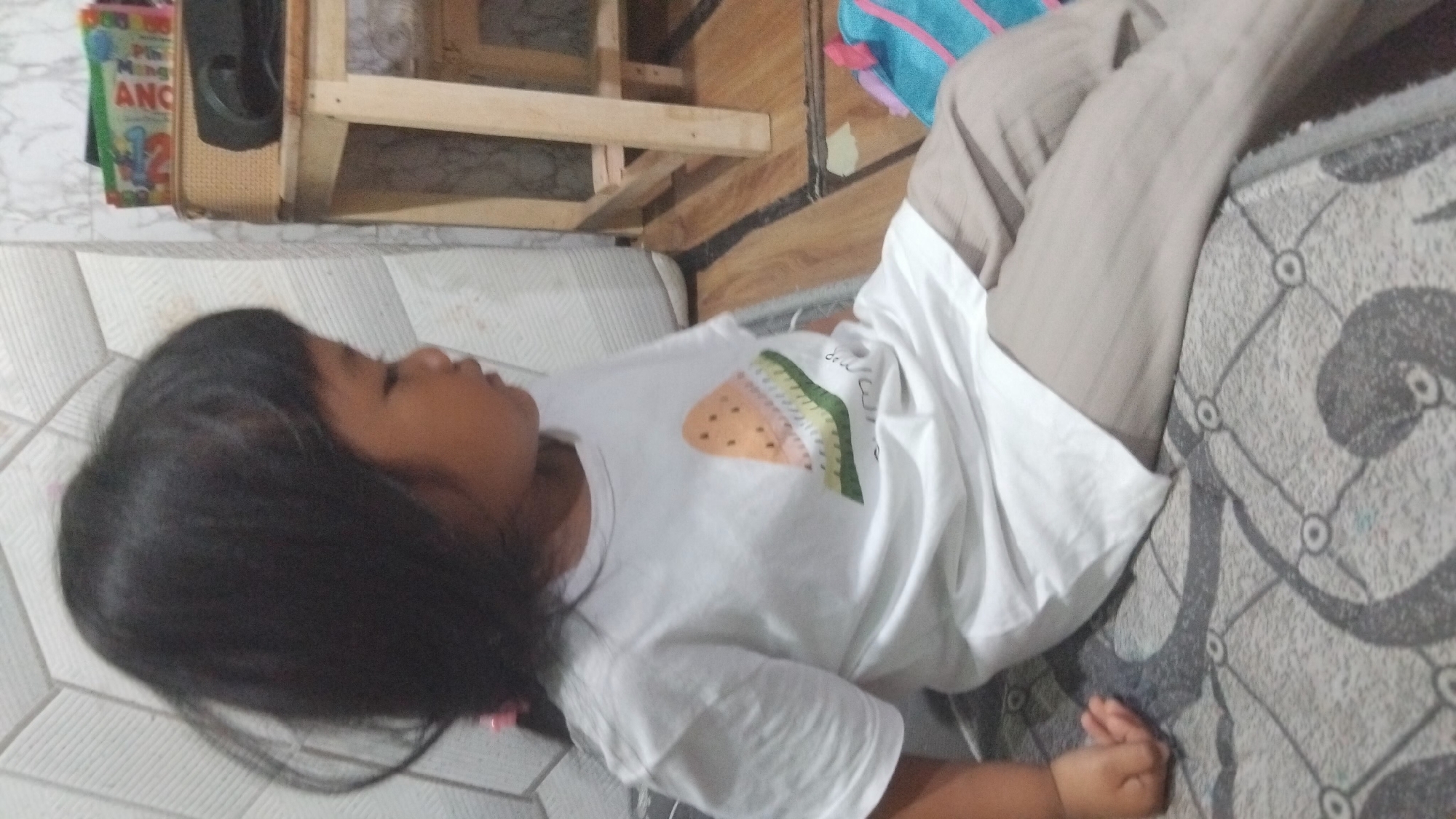 A young child with dark hair is sitting on a patterned mat, wearing a white t-shirt with a watermelon slice graphic and light brown striped pants. The child is facing to the right, with their head turned slightly. In the background, a wooden table with a woven basket and books is visible. A portion of a colorful backpack is also present on the right side of the image. The environment appears to be indoors, with tiled walls and wooden flooring. The setting is a residential interior in Bangun Rejo, Indonesia.