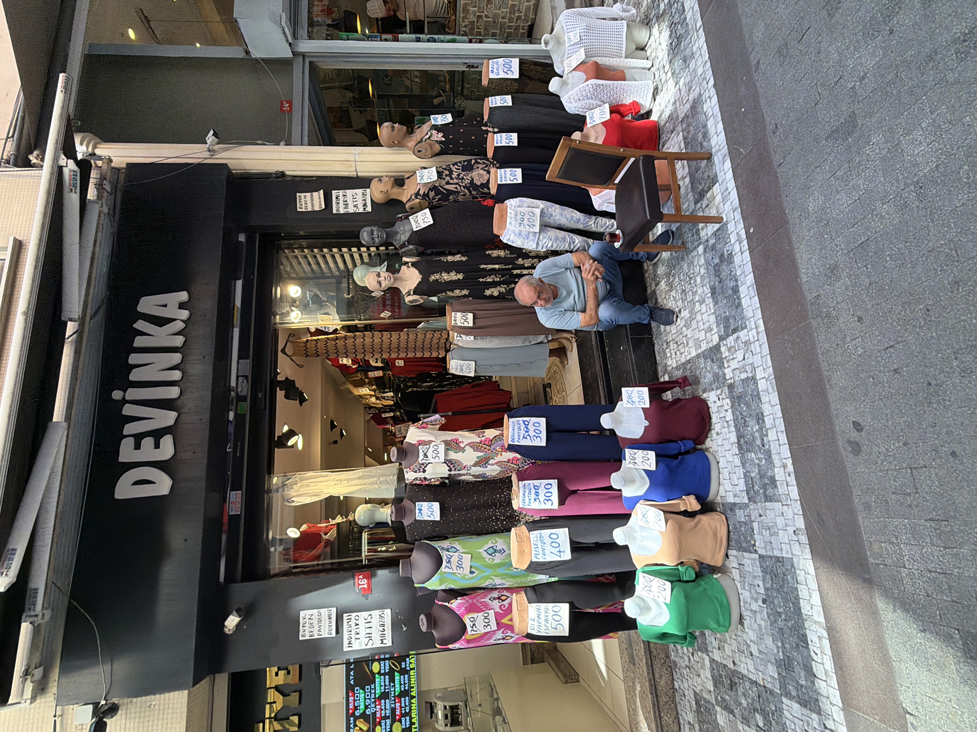 The image depicts a street-level view of a clothing store facade in Kadıköy, Turkey. The store, named "DEVİNKA," features a black awning above the entrance. In the storefront display, several mannequins are showcased, some wearing dresses and others displaying trousers and tops. Prices are displayed on signs in front of the mannequins. A man is seated on the sidewalk to the left of the entrance, facing the store and appearing to be resting. A chair is placed near him, and a small glass of tea is on the ground next to it. The street is paved with a patterned tile design. The overall scene suggests a typical commercial street in a Turkish city.