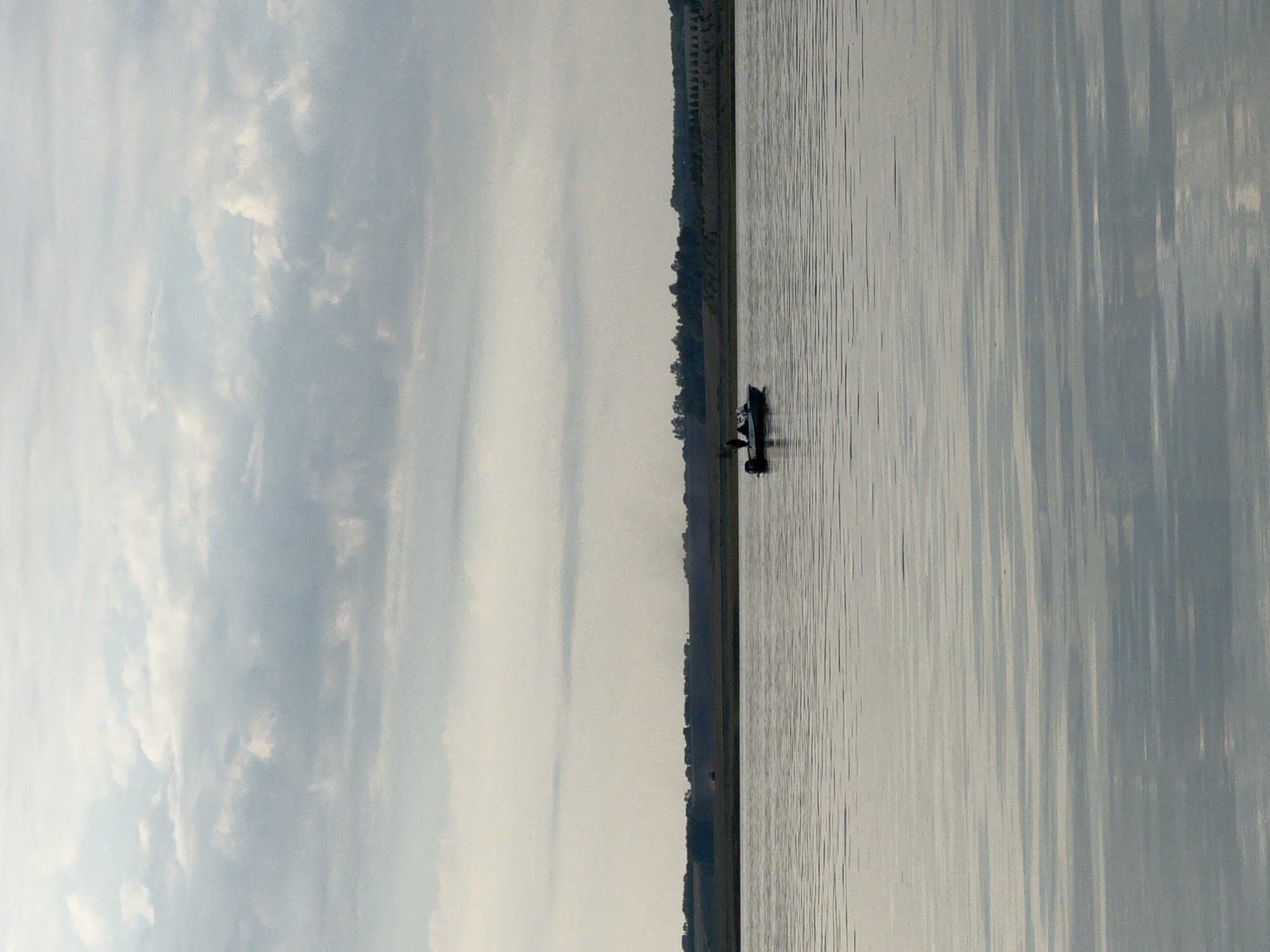 A medium shot captures a serene lake scene under a cloudy sky. In the foreground, the water's surface is calm, reflecting the muted tones of the sky and the distant shoreline. Small, horizontal ripples are visible, creating a textured pattern. A small motorboat with two people on board is situated in the middle ground, centered in the frame. The boat appears to be engaged in fishing, with the individuals potentially casting lines. The background features a tree-lined shoreline, with a gentle slope of cultivated fields or grassy hills behind it. The overall atmosphere is tranquil, with a hint of activity from the boaters. The location is the city of Novopichugovo, Russia.