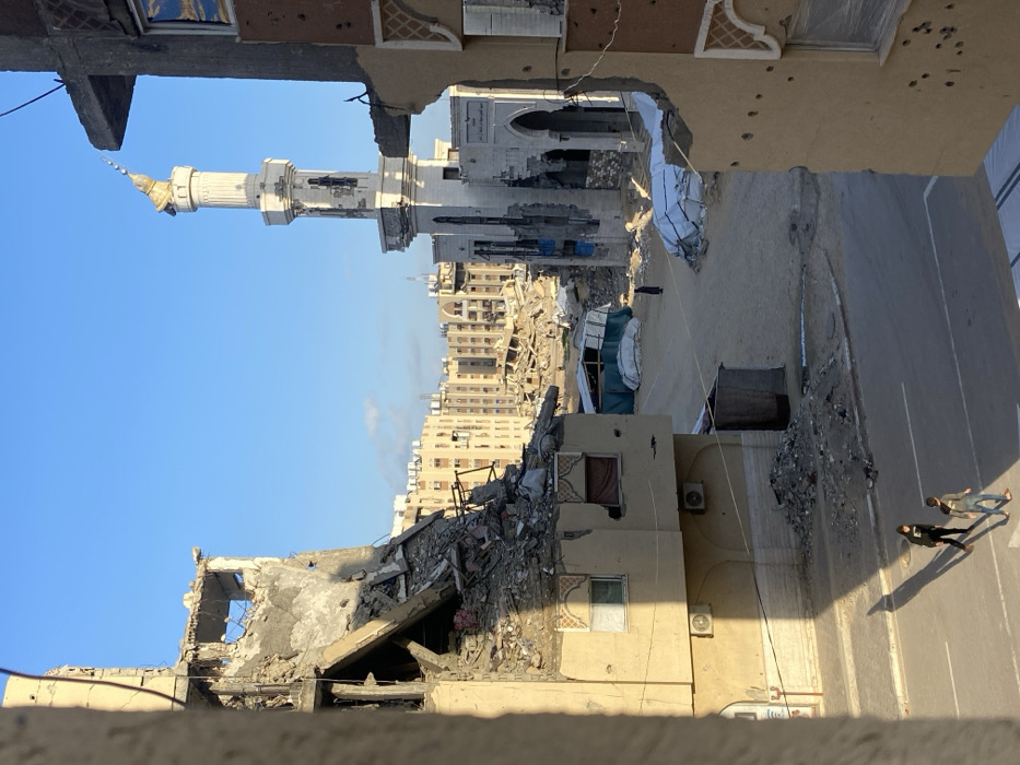 The image, appearing to be rotated 90 degrees clockwise from its natural orientation, depicts a scene of extensive urban destruction, viewed from an elevated perspective, likely from an upper floor of a building. The sky is clear blue, suggesting daytime.
In the foreground, the edge of a building or structure is visible at the very bottom, and immediately above it to the left, there's a severely damaged, grey-colored building with large sections of its walls and roof collapsed, exposing rebar and rubble. The remaining structure shows significant distress and holes. To the right of this collapsed building, another light-colored building stands, also showing signs of damage, including what appear to be bullet holes on its exterior wall. An air conditioning unit is mounted on its side.
Moving into the midground, a street or paved area runs diagonally from the bottom right towards the center, where it appears to be covered in debris. On the right side of this street, two or possibly three individuals are visible, casting long shadows, indicating a low sun angle, possibly late afternoon.
The midground and background reveal a broader landscape of devastation. On the left side, beyond the immediately visible rubble, more extensively damaged buildings are piled with debris. Further back, a prominent mosque with a tall, white minaret stands out against the blue sky. The very top spire of the minaret appears to be bent or partially collapsed. The main mosque building behind the minaret also shows signs of damage.
Beyond the mosque and the immediate foreground structures, numerous multi-story residential buildings extend into the distance. Many of these buildings, particularly those closer to the central damaged area, appear to be partially or completely destroyed, reduced to rubble, or severely damaged. Further in the distance, a faint plume of smoke or haze rises above the cityscape on the right. Several makeshift shelters or tents, constructed from white and green tarpaulins, are visible amidst the rubble in the central-right portion of the image. The overall impression is one of widespread conflict-related destruction and urban decay.