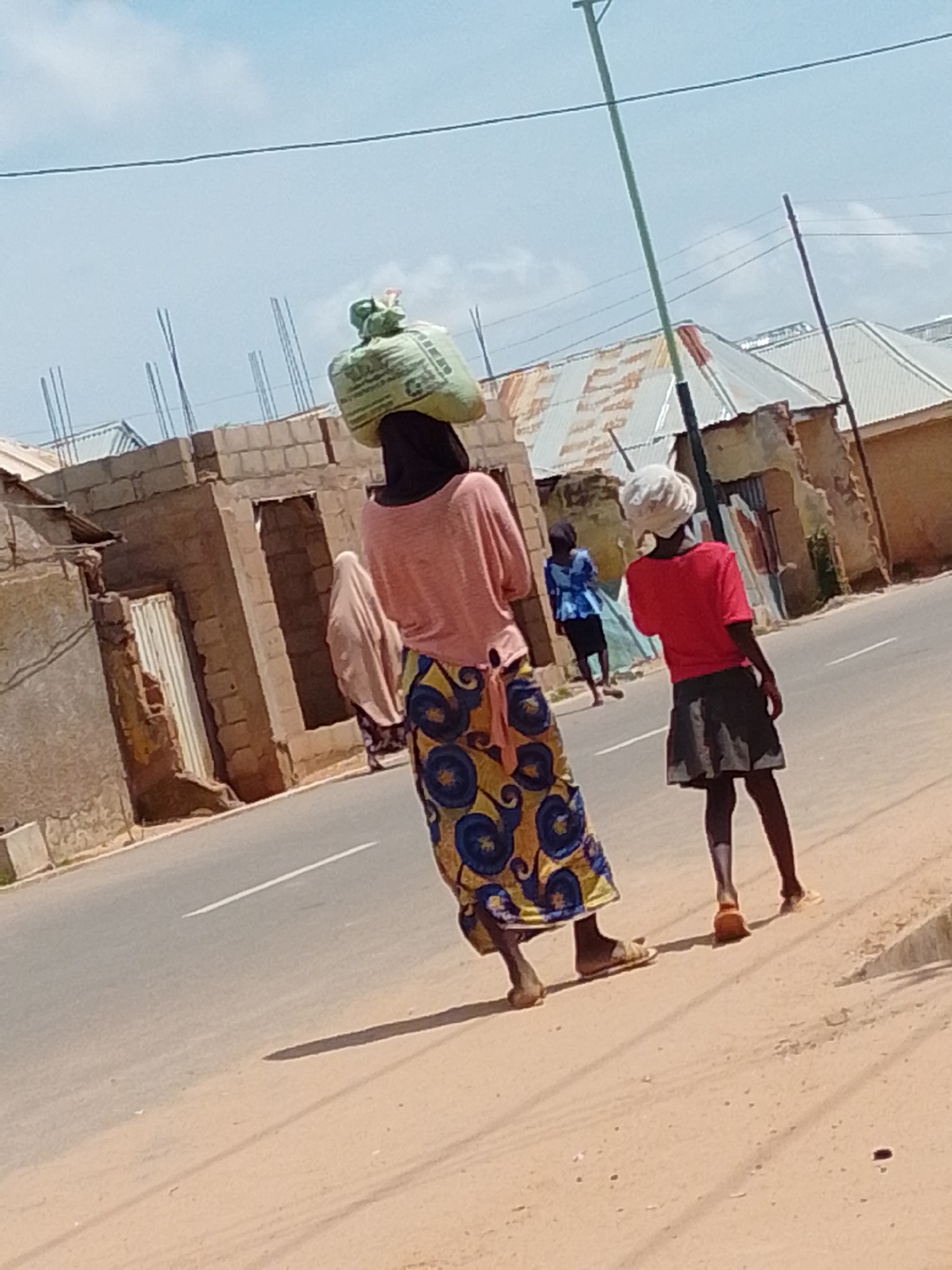 Two women and a child are walking on a dusty road in an urban environment. The woman in the foreground on the left is carrying a large, green bag on her head. She is wearing a pink shirt and a long, colorful skirt with a blue and yellow pattern. The woman in the middle appears to be wearing a light brown headscarf and a patterned dress. The child on the right is wearing a red shirt and a dark skirt, with a white headscarf. They are walking past buildings with unfinished brickwork and corrugated metal roofs. The sky is partly cloudy and blue. The road is paved with a white line marking the lane.