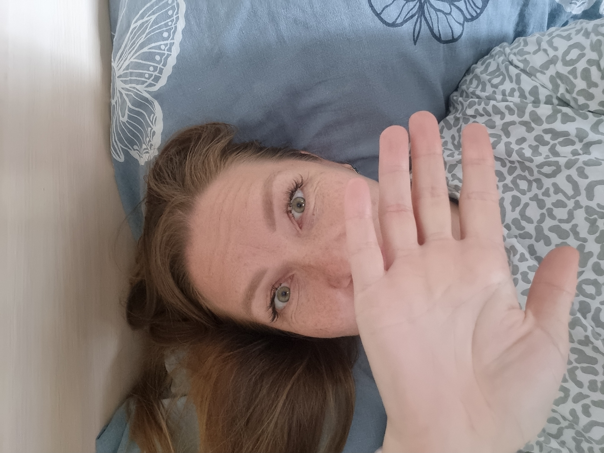 A woman with auburn hair and green eyes lies in bed, her right hand partially obscuring her face. She has freckles across her cheeks and nose. She is wearing a gray and white patterned top. The bedding is blue with a white butterfly pattern. The image is taken from a high angle, looking down at the woman. The environment is consistent with a bedroom in Chaikovsky, Russia.