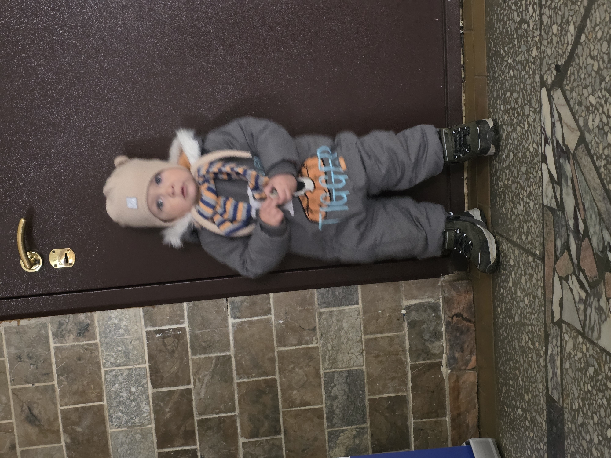 A baby wearing a gray snowsuit with an orange tiger print and a beige hat with bear ears lies on a dark brown surface next to a brick floor and a marble-patterned floor. The baby is holding an object with both hands. A brass door handle and lock are visible on the brown surface. The location is Chaikovsky, Russia.