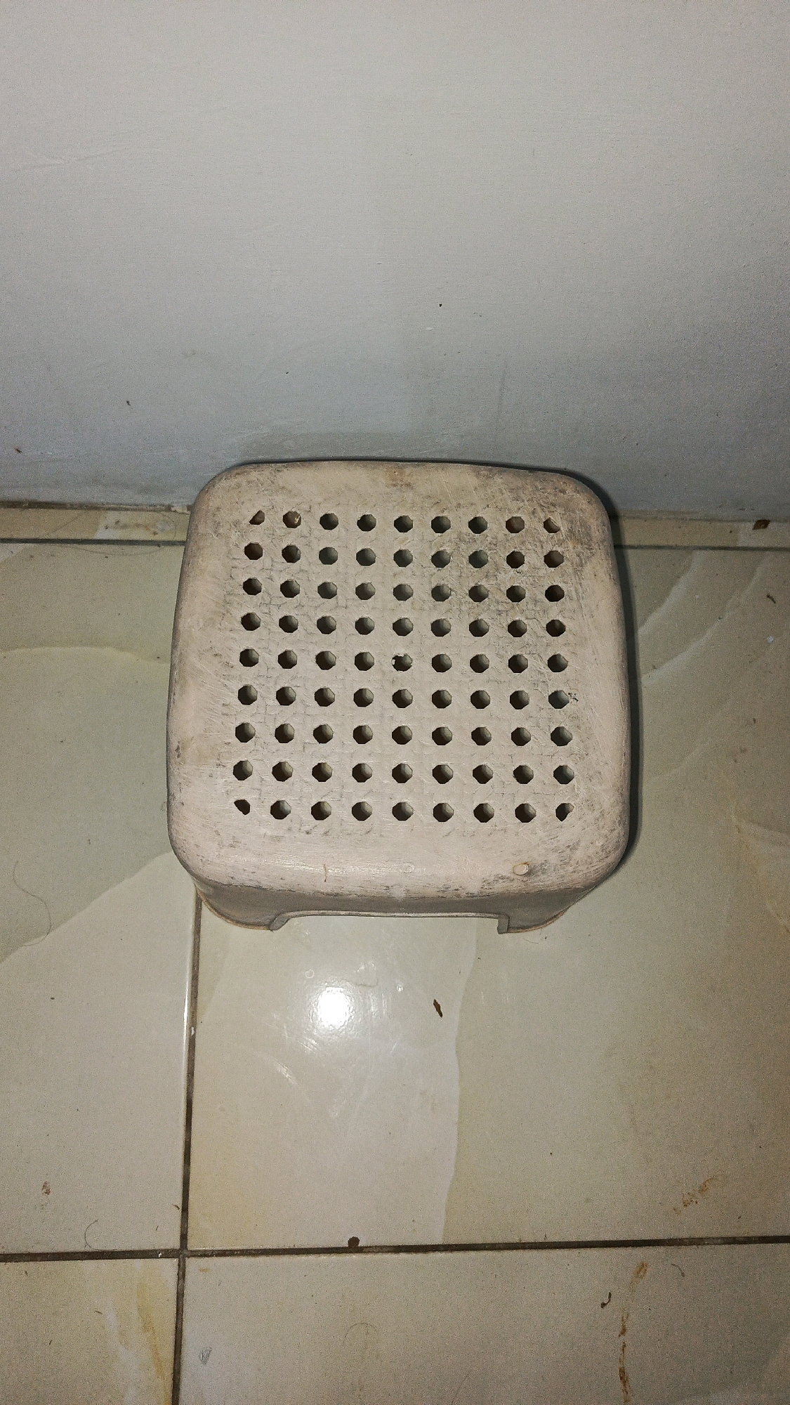 A square, off-white plastic stool with a perforated top sits on a tiled floor. The stool is slightly worn and dusty. The floor tiles are light-colored with visible grout lines. A wall, also off-white, is visible in the background above the stool. The lighting in the scene is dim, casting shadows on the floor. The image was taken in Babadan, Indonesia.