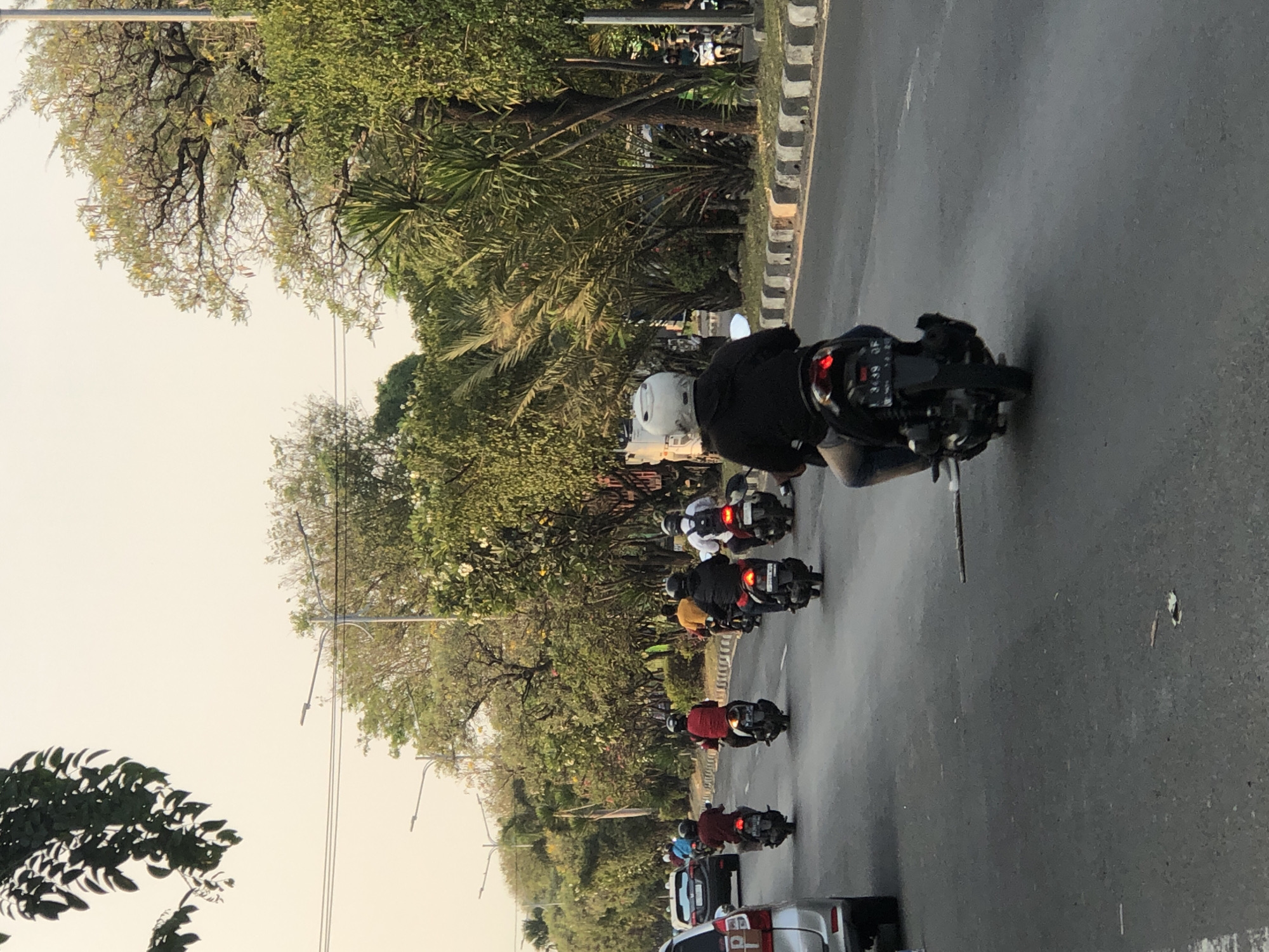 The image captures a street scene in Ngaban, Indonesia, on a clear day with a pale yellow sky. Several motorcycles are traveling along an asphalt road, with the majority moving away from the viewer. A person in the foreground is riding a black motorcycle, wearing a white helmet and a dark jacket. To the left, a silver car is visible. The road is lined with trees, creating a green canopy along the side. Streetlights are also present along the road. Several other motorcycles with riders, some wearing helmets and brightly colored shirts, are seen further down the road. The overall impression is of a moderately busy street in an urban environment with abundant greenery.