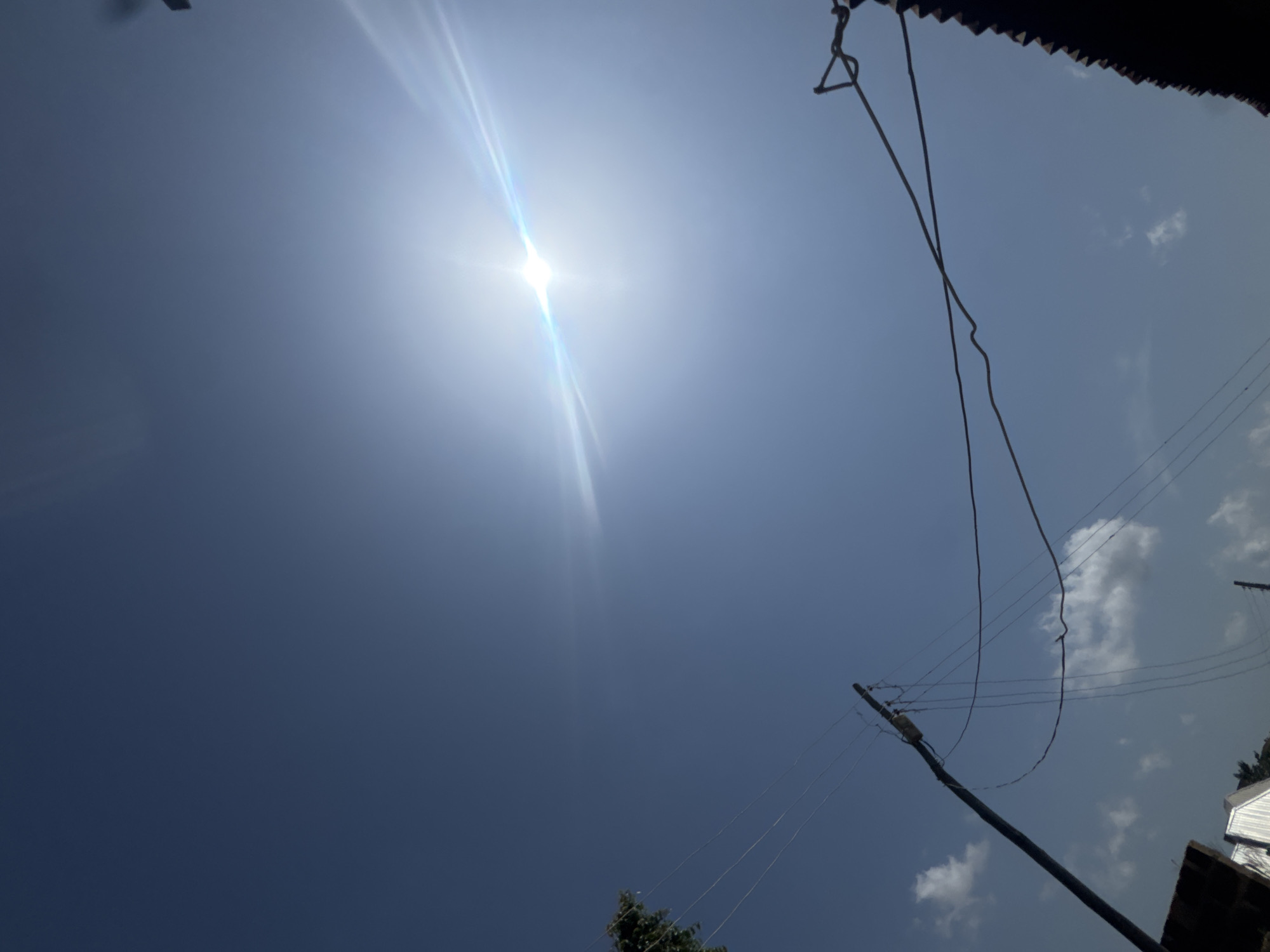 The image shows a bright sun in a clear blue sky, with some light clouds visible. In the foreground, a wooden utility pole with electrical wires is prominent. The pole is leaning to the left, and several wires are strung between it and other unseen points, some of which are sagging. A dark, textured object, possibly a roof with jagged edges, is visible on the right side of the frame. On the left side, a portion of a tree with green foliage is seen, along with the corner of a building and a white structure that appears to be part of a house. The scene suggests an outdoor setting, possibly in a residential area, under a sunny sky.