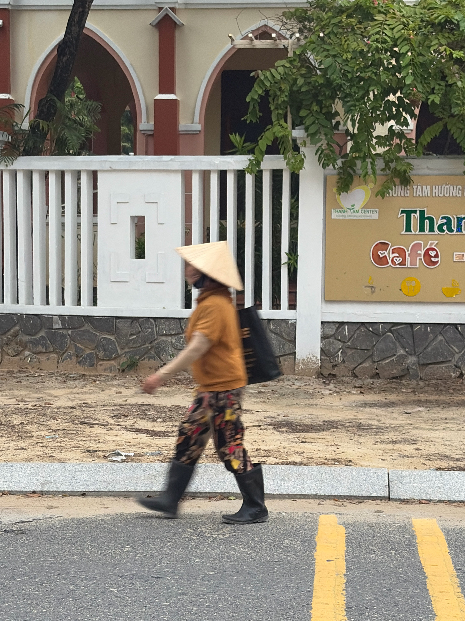 A person wearing a conical hat, a tan shirt, patterned pants, and black boots walks across a street in Da Nang, Vietnam. The person carries a black bag over their shoulder. Behind the person is a white fence with a stone base, a sandy area, and a building with arched windows. A sign for "THanh TÁM CENTER" and "Cafe" is visible on the building. Yellow lines mark the street.