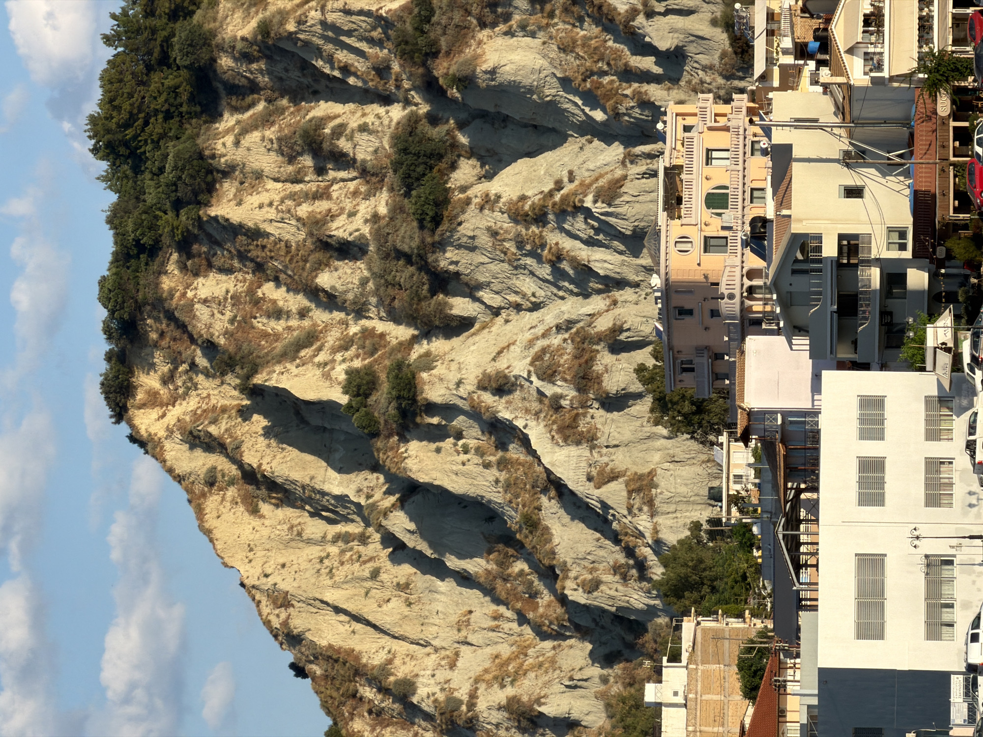 A sunlit, medium-shot photo captures the town of Zakynthos, Greece, nestled at the base of a towering, eroded hillside. The hillside, with its steep, striated slopes of pale earth, is sparsely covered with dry grasses and a few scattered trees. In the foreground, several buildings of varying heights and architectural styles are visible. A prominent, modern, white building with horizontal window louvers occupies the left side of the frame. To its right, a collection of multi-story buildings, some painted in warm tones like peach and cream, feature balconies, windows, and visible rooftops. The sky above is a clear blue with a few wispy white clouds. The overall scene suggests a coastal town in a Mediterranean climate, with the natural landscape dominating the background and urban development in the foreground.