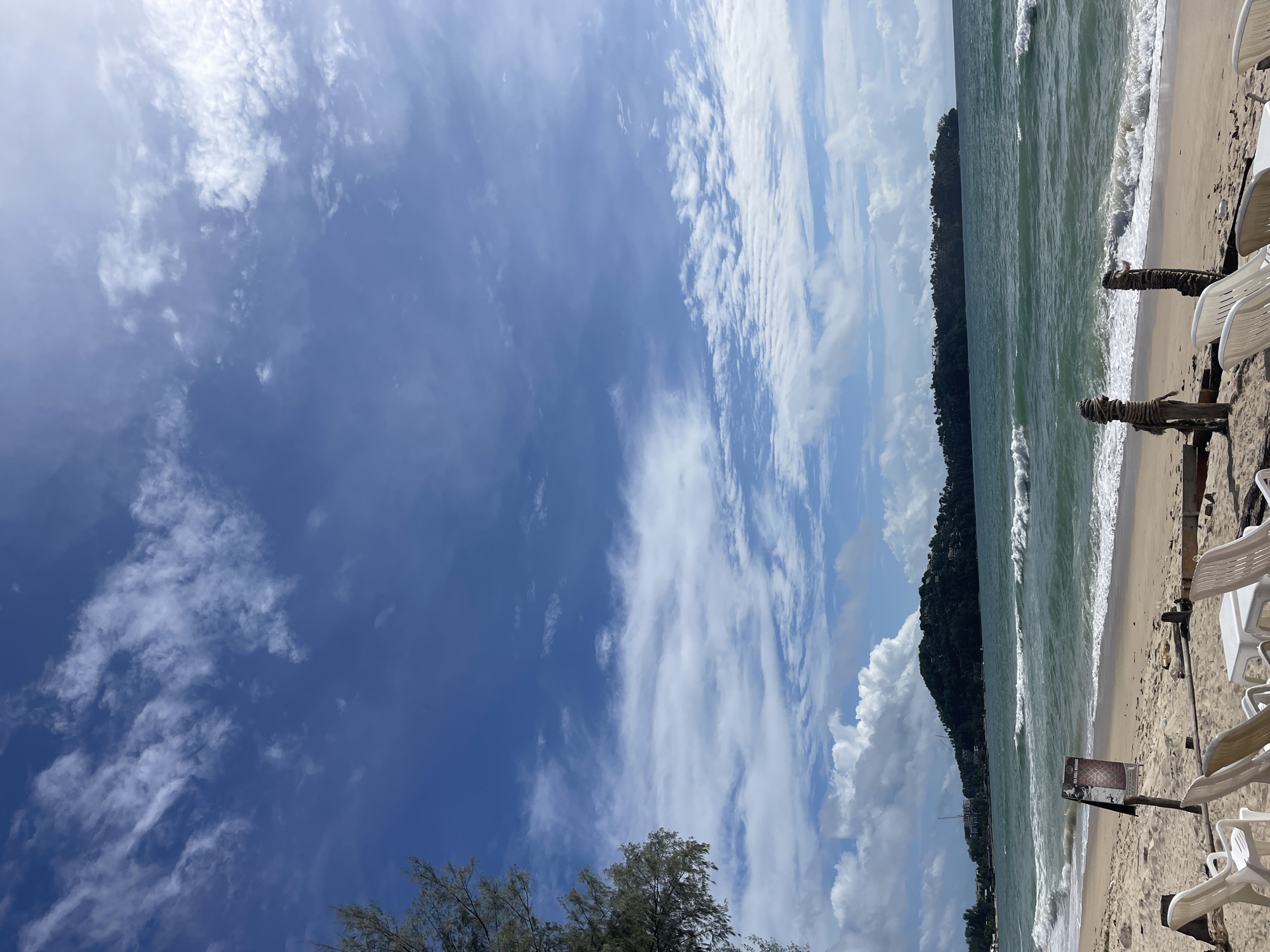 An eye-level, medium shot captures a tranquil beach scene in Choeng Thale, Thailand, under a vast, blue sky adorned with scattered white clouds. The sandy shore stretches across the foreground, dotted with several empty white plastic lounge chairs arranged in neat rows. A small, white plastic table is positioned between some of the chairs. In the mid-ground, gentle waves with white crests roll onto the shore, creating a serene oceanfront. A dark sign with white text is mounted on a wooden pole near the water's edge. In the distance, a lush, green, tree-covered island rises from the turquoise sea, with a few buildings visible on its slopes. The horizon is clear, with the ocean meeting the sky under the bright daylight.