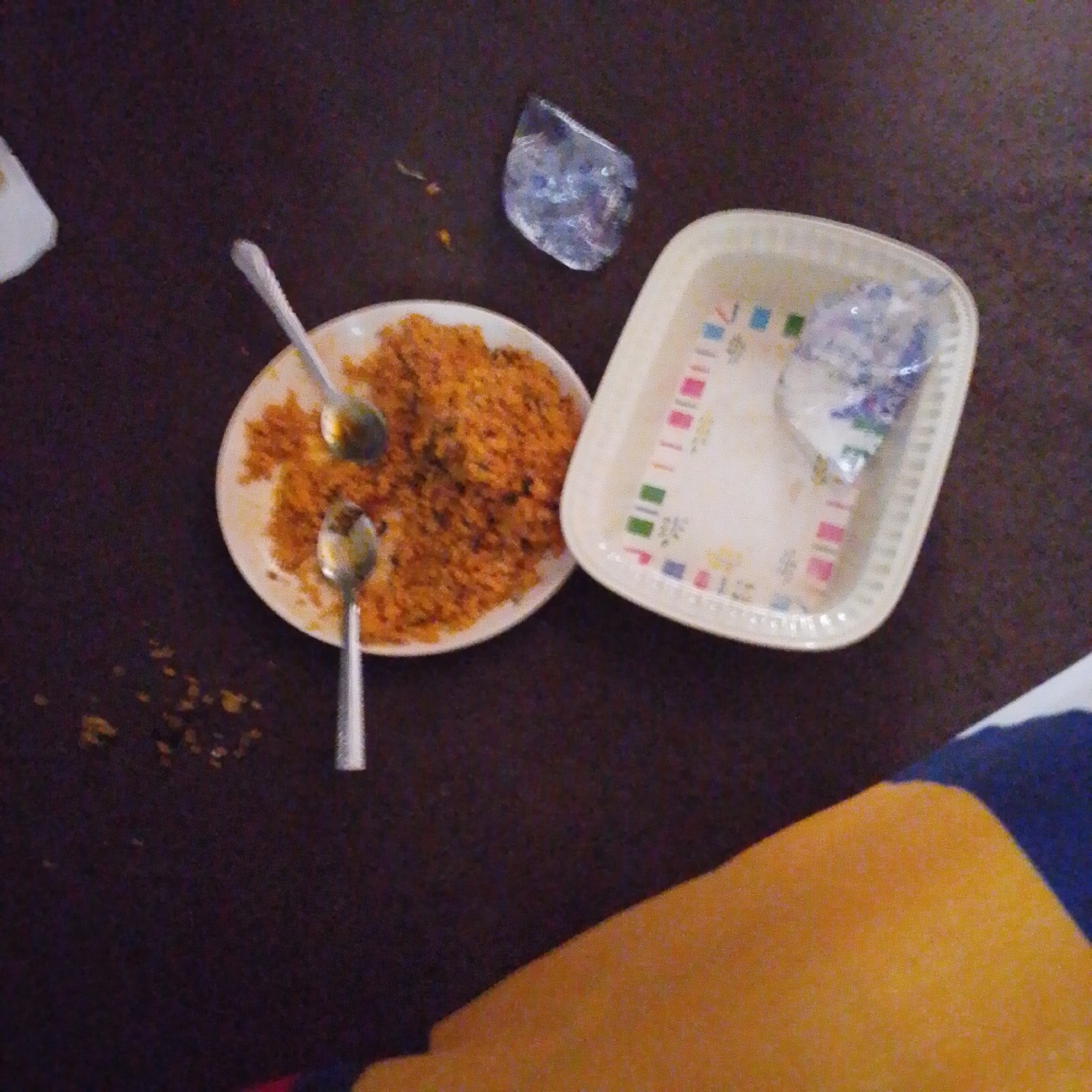 The image shows a close-up, overhead view of a table with food and utensils. In the center, a white plate contains a serving of rice mixed with what appears to be spices, giving it an orange hue. Two metal spoons are resting in the plate, one slightly to the left and another further down. To the right of the plate is a white plastic tray with a colorful decorative pattern along the rim. Inside the tray, there's a crumpled piece of plastic, possibly packaging or a napkin. To the upper right of the plate, a small, irregular piece of clear material, possibly plastic or ice, is visible on the dark brown surface of the table. Scattered on the table around the plate are a few small, unidentifiable dark specks. In the lower right corner of the image, a portion of what appears to be a person's clothing, with a yellow and blue pattern, is visible, suggesting the presence of someone at the table. The overall lighting is dim, and the image quality is grainy, typical of indoor or low-light photography. The scene depicts remnants of a meal, likely in a casual dining setting, possibly at a home or a small eatery in Jalingo, Nigeria.