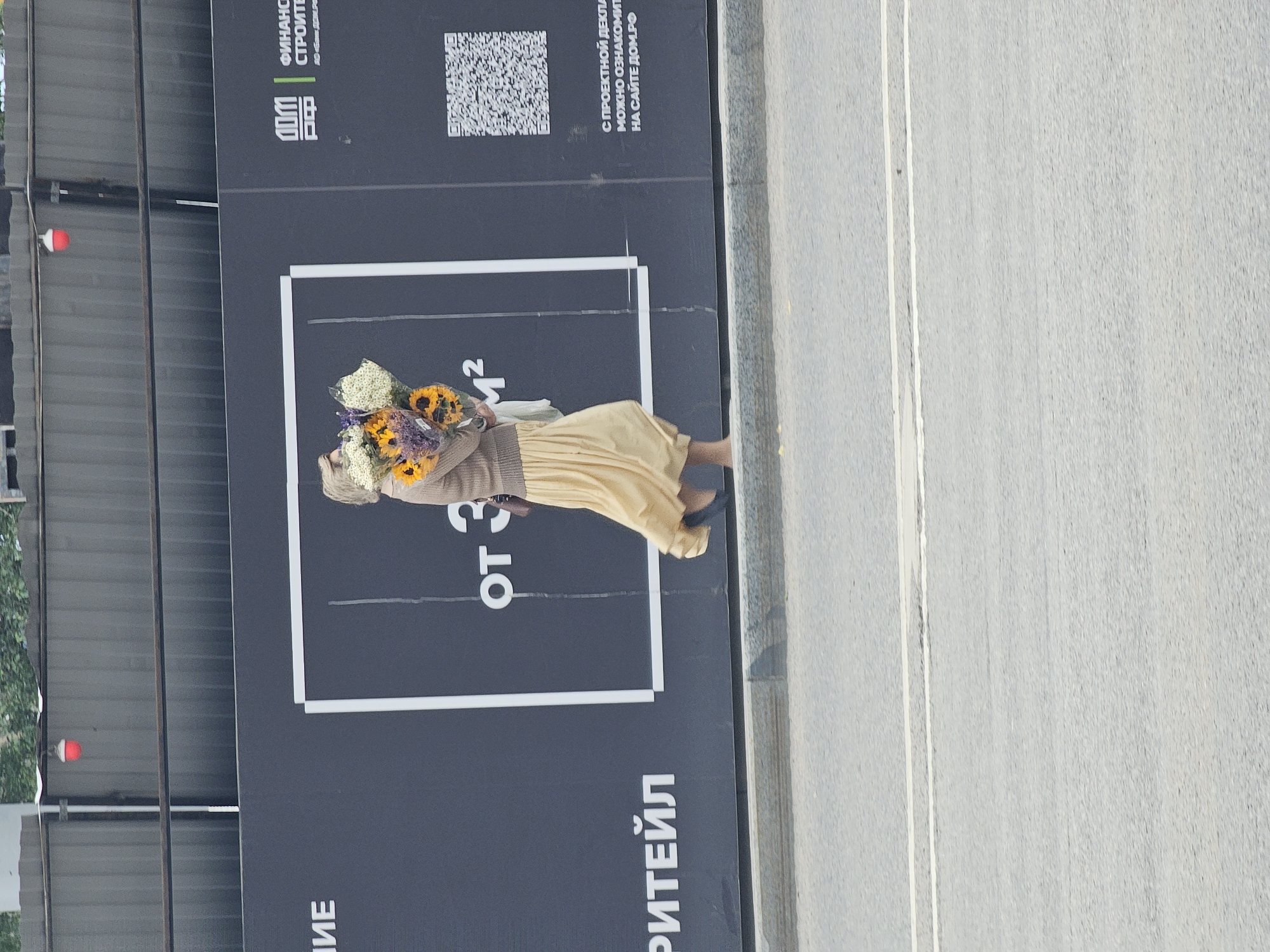 This media file shows a person, likely a woman, holding a bouquet of sunflowers and other flowers. She is standing in front of a large dark billboard that displays text and a QR code. The billboard appears to be an advertisement for a real estate development, with text in Russian indicating "FROM 3" and mentioning "PROJECT DECLARATION" and a website "DOM.RF". The scene is outdoors, with a grey asphalt road visible to the right of the billboard, suggesting an urban setting. The lighting indicates it is daytime. The woman is wearing a light-colored skirt and a brown cardigan. The overall impression is that of a marketing event or a staged photo opportunity related to the advertised property.