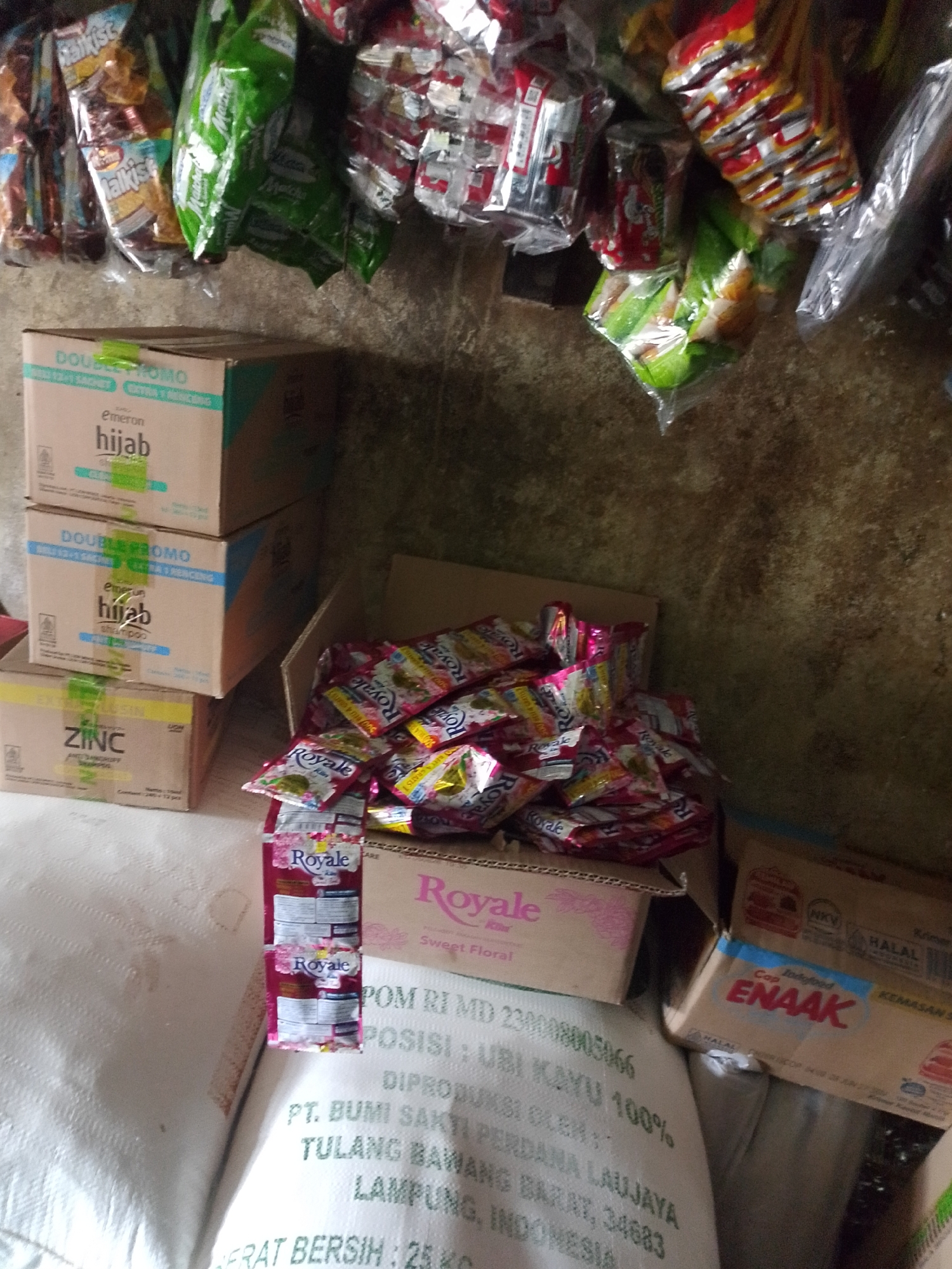 This is a close-up, high-angle, eye-level shot of a shelf display in a small shop or warung, likely located in Palembang, Indonesia. The overall impression is one of a cluttered, somewhat disorganized retail environment.
In the foreground, a large white sack, possibly containing rice or flour, takes up the left side of the frame. It has green text printed on it, which is partially visible and appears to be product information or branding.
On the right side of the image, various products are stacked and displayed. There are several boxes of shampoo, identifiable by the brands "Emeron Hijab Shampoo" and "ZINC Anti Dandruff." Above these boxes, a collection of packaged snacks or candies in colorful wrappers are hanging or loosely piled.
Further to the right, a cardboard box is filled with small packets, predominantly pink and yellow, bearing the brand "Royale." The packets appear to be related to laundry detergent or fabric softener. Another box with red and blue labeling, featuring the brand "Indofood Cap ENAAK," is also visible, suggesting it might contain food items.
The background consists of a textured, likely concrete or stucco wall with a mottled, greenish-brown appearance, indicating age or possibly dampness. The lighting appears to be artificial, creating some shadows and highlighting the textures of the products and the wall. The scene suggests a typical small-scale retail operation where everyday consumer goods are sold.