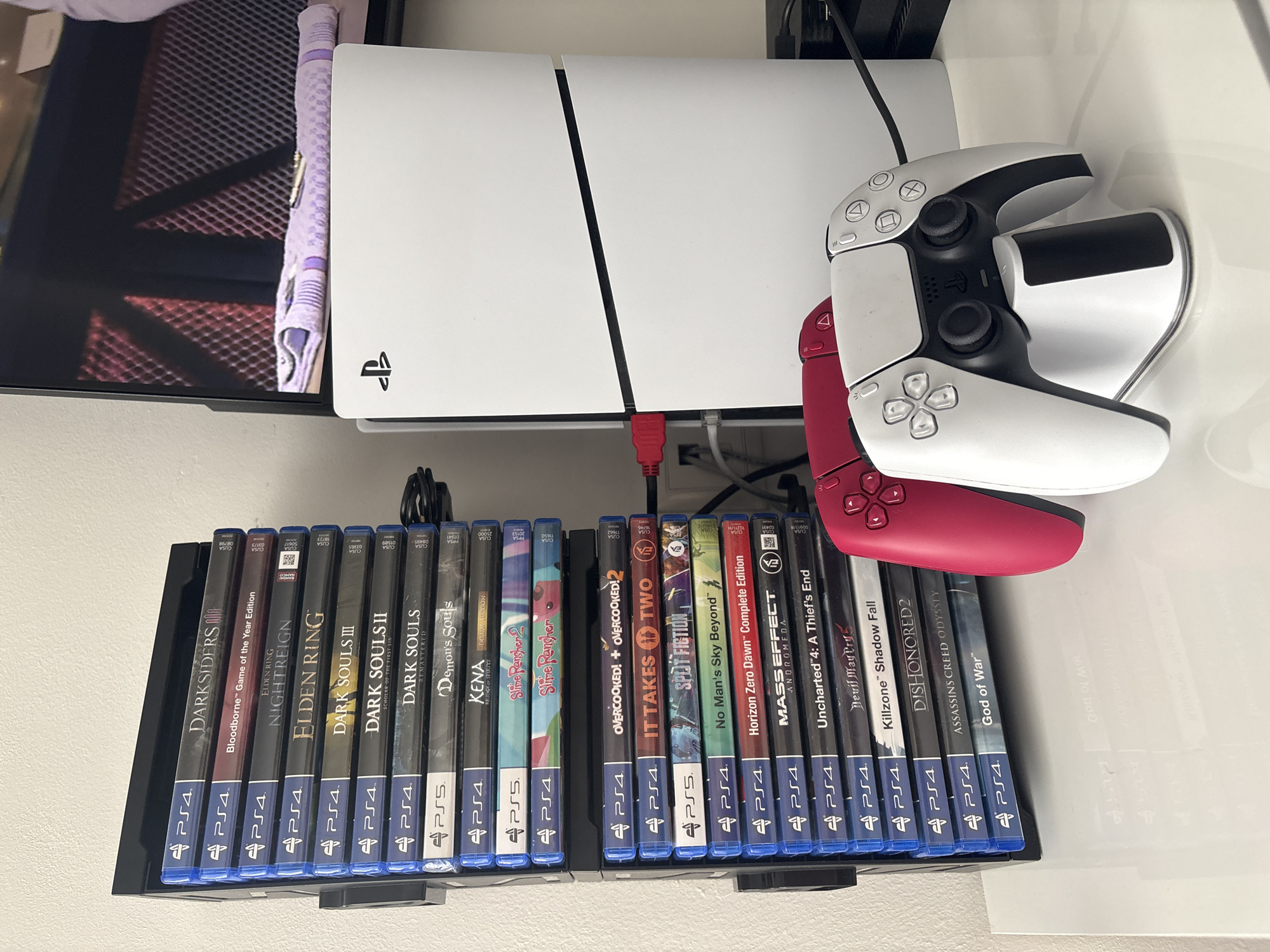 This image displays a gaming setup located in Plovdiv, Bulgaria, featuring a PlayStation 5 console, two DualSense controllers, and an extensive collection of PlayStation 4 and PlayStation 5 game cases.
The white PlayStation 5 console is positioned vertically on a clean, white surface, likely a desk or shelf. A red HDMI cable is visibly connected to the console's rear, leading towards a partially visible television screen on the left, which shows a dark, patterned image.
Adjacent to the console are two PlayStation DualSense controllers. One is white, matching the console's aesthetic, while the other is a distinct red. Both controllers are neatly placed on a stand or charging dock, appearing ready for use.
Below the main console setup, a black shelf holds a substantial library of video game cases, organized into two distinct rows. The majority of the titles are for the PlayStation 4, interspersed with several PlayStation 5 games. Visible titles include "Darksiders III," "Bloodborne Game of the Year Edition," "Elden Ring," "Dark Souls III," "Dark Souls Remastered," "Demon's Souls" (PS5), "Kena Bridge of Spirits" (PS5), "Slime Rancher 2" (PS5), "Overcooked! + Overcooked! 2," "It Takes Two," "No Man's Sky Beyond," "Horizon Zero Dawn Complete Edition," "Mass Effect Andromeda," "Uncharted 4: A Thief's End," "Devil May Cry 5," "Killzone Shadow Fall," "Dishonored 2," "Assassin's Creed Odyssey," and "God of War." The collection showcases a variety of genres, with a notable presence of action-adventure and role-playing games. The overall scene suggests a well-maintained and organized gaming enthusiast's area.