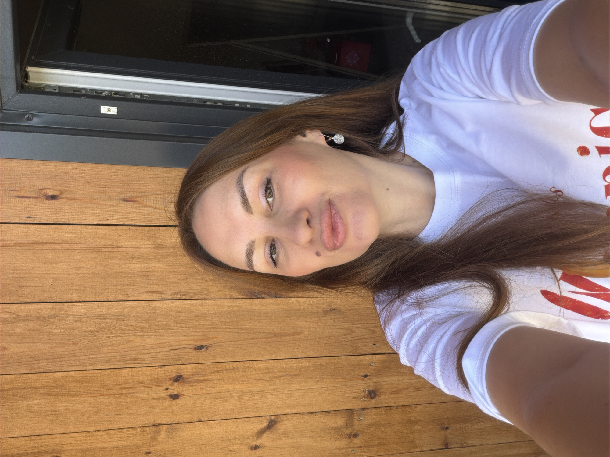 A woman with long, brown hair and fair skin is taking a selfie in front of a wooden wall. She is wearing a white t-shirt with red lettering. She has on a sparkly earring in her right ear. Her lips are slightly pursed, and she has a mole above her upper lip on the left side. The background shows a wooden wall and a black doorway. The lighting suggests it is daytime. The image is taken from the chest up.