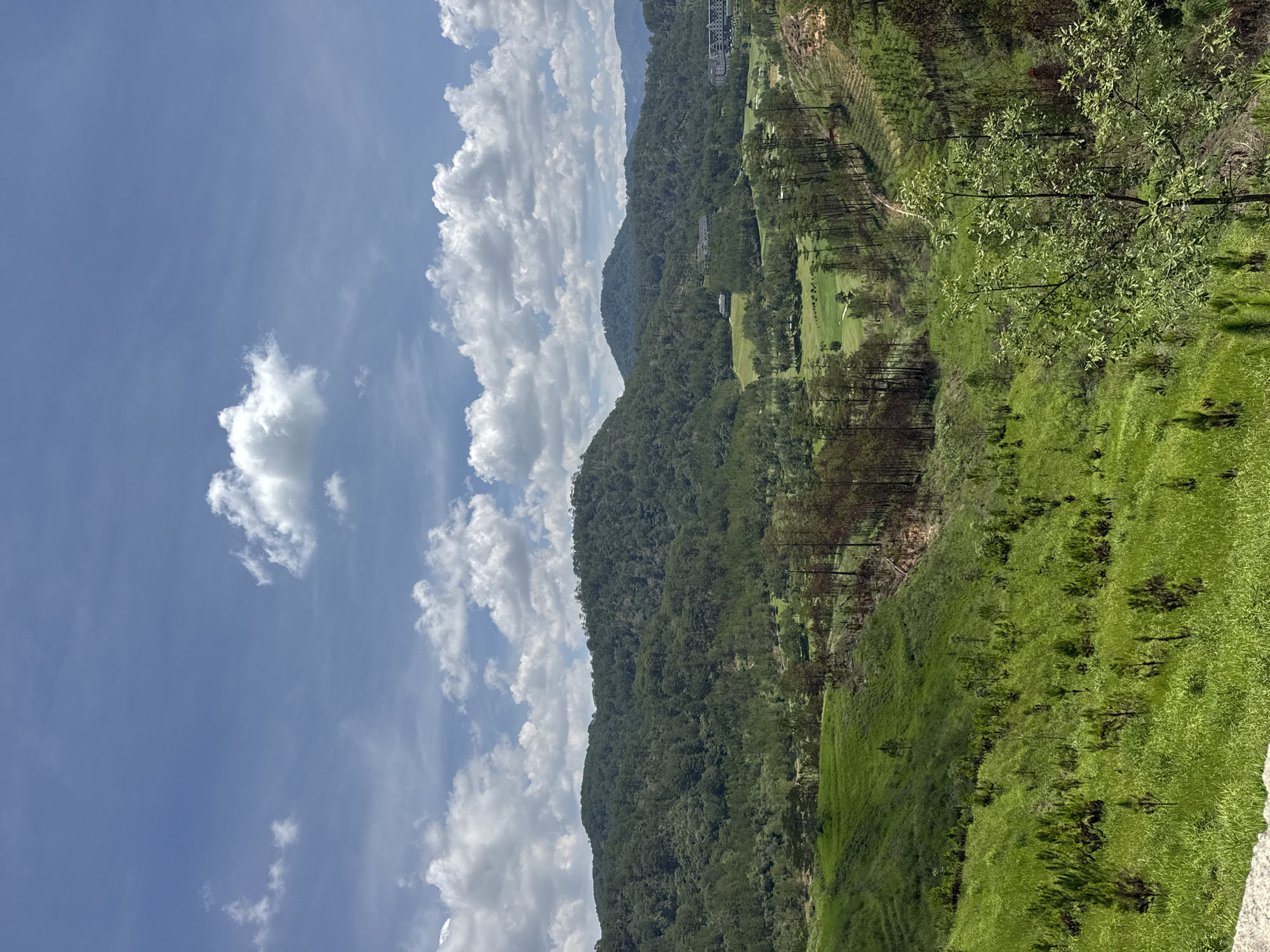 The image presents a view of a mountainous landscape under a partly cloudy sky. Green foliage covers the terrain, suggestive of lush vegetation. A section resembling a golf course is visible among the trees. A building structure is situated further up the mountain. The location is likely in a region characterized by hilly terrain and a temperate climate suitable for dense vegetation. The image is not located in the city of Hiep Thanh, Vietnam.