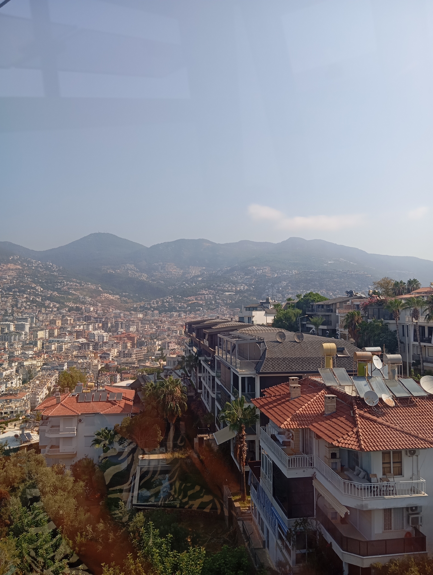 The image is an aerial view of a densely populated city nestled against a backdrop of rolling, hazy mountains under a clear sky. Buildings, mostly residential with red-tiled roofs, occupy the landscape, extending up the slopes of the hills. In the foreground, a cluster of modern multi-story buildings with balconies and solar water heaters are visible. Lush green trees and foliage surround some of the buildings. A swimming pool with a camouflage pattern is visible in the lower right quadrant. The scene suggests a warm, sunny day in a coastal or mountainous urban environment.