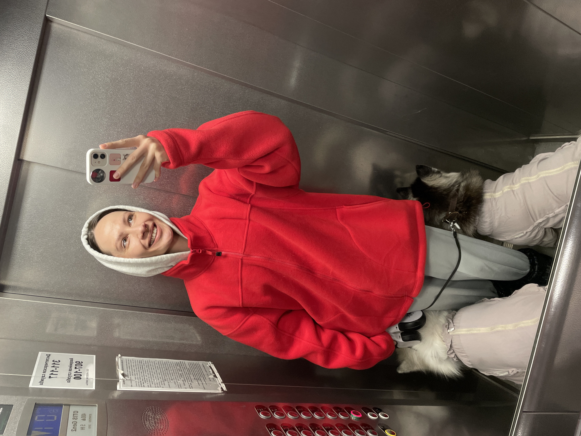 A smiling woman takes a selfie in an elevator. She wears a red fleece jacket with a gray hood underneath. She holds a white smartphone decorated with stickers. Beside her are two dogs, one black and white and one white, both wearing light-colored coats. The elevator has silver walls, a panel with red buttons, and a notice in Russian, "Правила пользования лифтом" and "301-100" (Rules for using the elevator and 301-100). The image was taken inside a building, presumably located in Izhevsk, Russia, based on the Russian text on the notice.