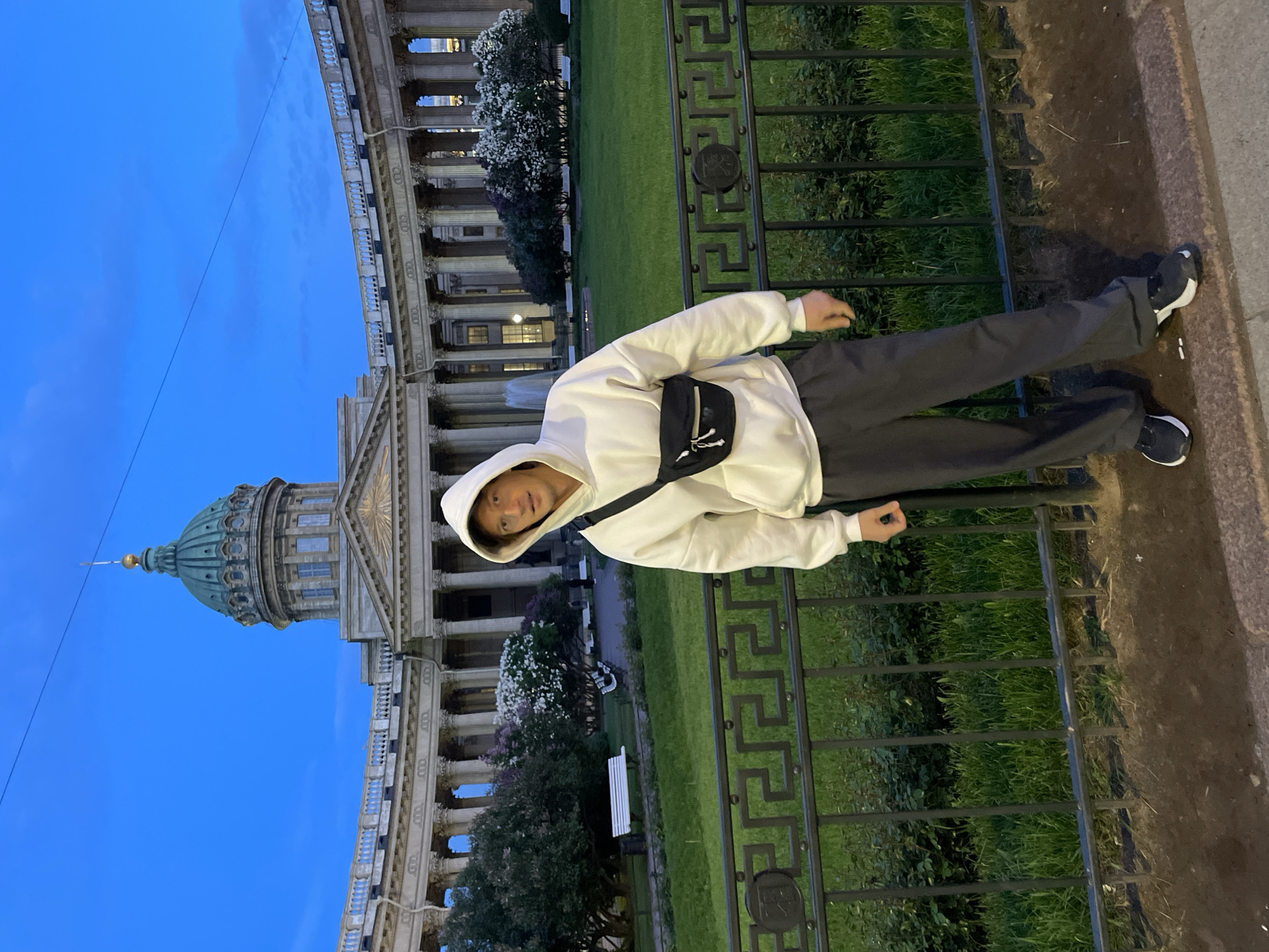 A young man is featured in a full-body outdoor shot, standing beside a black, ornate metal fence. He is wearing a white hooded sweatshirt, dark trousers, and black sneakers with white soles. A black fanny pack or crossbody bag is worn across his chest. He looks towards the camera with a neutral expression.
Behind him, the distinctive Kazan Cathedral in Saint Petersburg, Russia, dominates the background. Its prominent green dome and the grand semi-circular colonnade with numerous columns are clearly visible. Beyond the fence, a well-maintained grassy area with trees and bushes, including some with white and purple flowers (likely lilac), stretches towards the cathedral. A white bench can also be seen in the distance on the lawn.
The sky is a deep blue, indicating that the photograph was taken during dusk or late evening. Some internal lights are visible within the windows of the cathedral, suggesting the transition from day to night. The overall scene captures the man against the backdrop of an iconic architectural landmark. There is no visible text in the image.