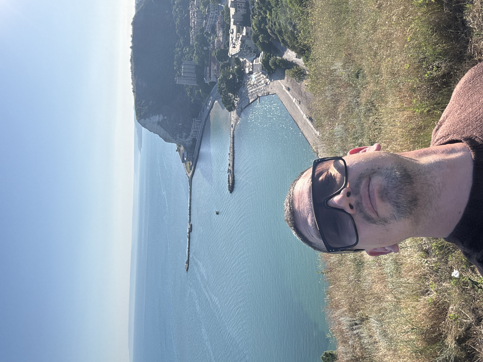 This is a selfie taken from a hillside overlooking a coastal town. The photographer, a man with short hair, sunglasses, and stubble, is in the foreground. He is wearing a brown knitted sweater. Behind him, the sea is a bright turquoise, with a few small boats visible. In the middle ground, there is a town built on the coast, with white buildings and green trees. The town is nestled against a steep, white cliff face. The sky is clear and blue. The area appears to be sunny and warm. The vantage point suggests the location is in the vicinity of Kavarna, Bulgaria, known for its dramatic coastal cliffs and proximity to the Black Sea.