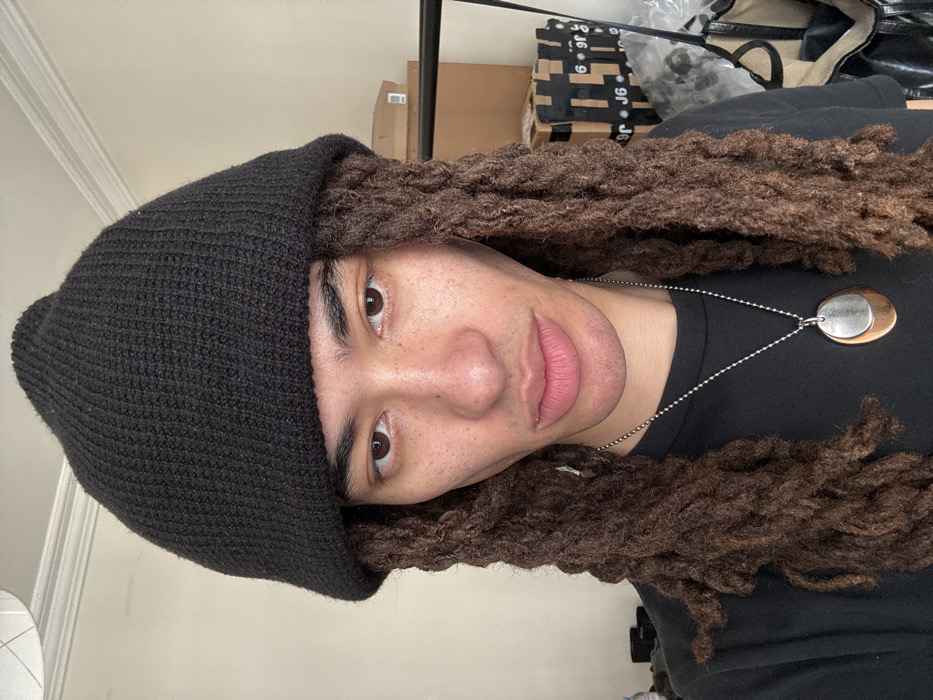 The image depicts an indoor scene featuring one individual, appearing to be male, facing the camera directly. The person has light-brown skin, visible freckles on the cheeks and nose, dark brown eyes, and dark brown dreadlocks that extend past the shoulders. They are wearing a black ribbed knit beanie, a plain black t-shirt, and a silver-colored ball chain necklace with a two-tone oval pendant (silver-colored layered over copper-colored). The individual maintains a neutral facial expression. The background features a plain light-colored wall on the left side of the frame. On the right side, several cardboard boxes are stacked, with one box displaying partially visible text including "6. J6" and "99. Paris". A black metal structure, possibly a clothing rack, is partially visible behind the individual's right shoulder, and a light-colored bag with dark trim is near the bottom right corner. The specific text "Paris" on a cardboard box confirms the location as Paris, France.