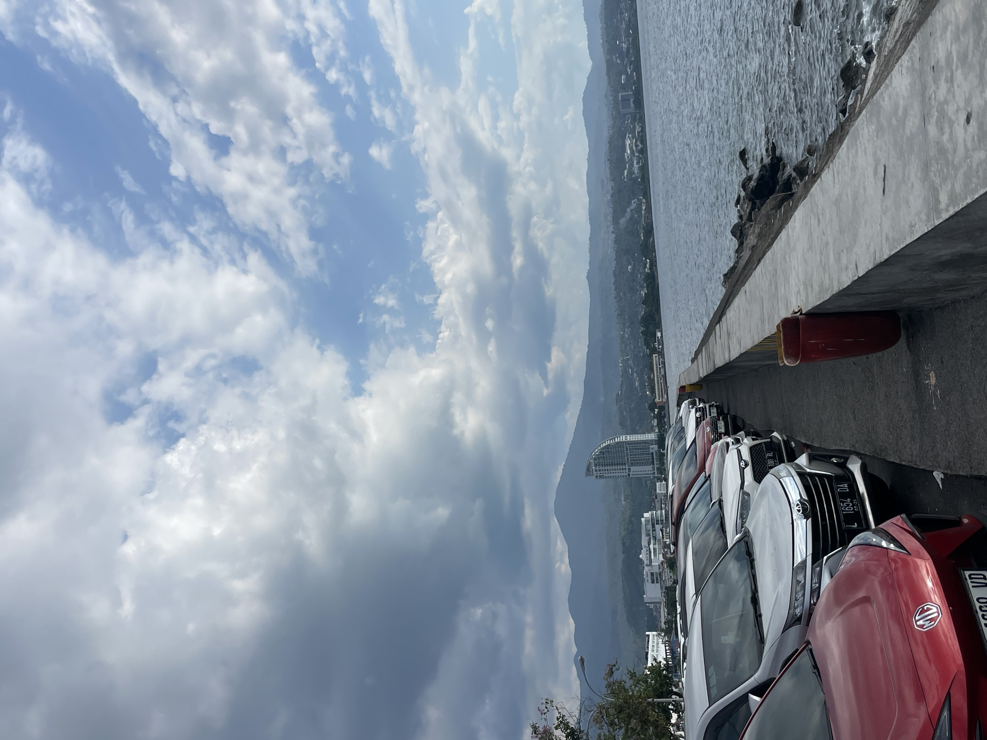 The image depicts a waterfront parking area in Manado, Indonesia, with a view of the city and surrounding hills. In the foreground, several cars are parked along a paved area next to a concrete seawall. The seawall is topped with a metal railing and a red trash can is visible. The water of the bay is calm, with small waves lapping against the shore. Beyond the parking area, buildings, including a distinctive modern high-rise, line the waterfront. In the background, rolling hills and mountains form a picturesque backdrop under a cloudy sky. The lighting suggests it is daytime, with the clouds creating a dramatic and varied sky.