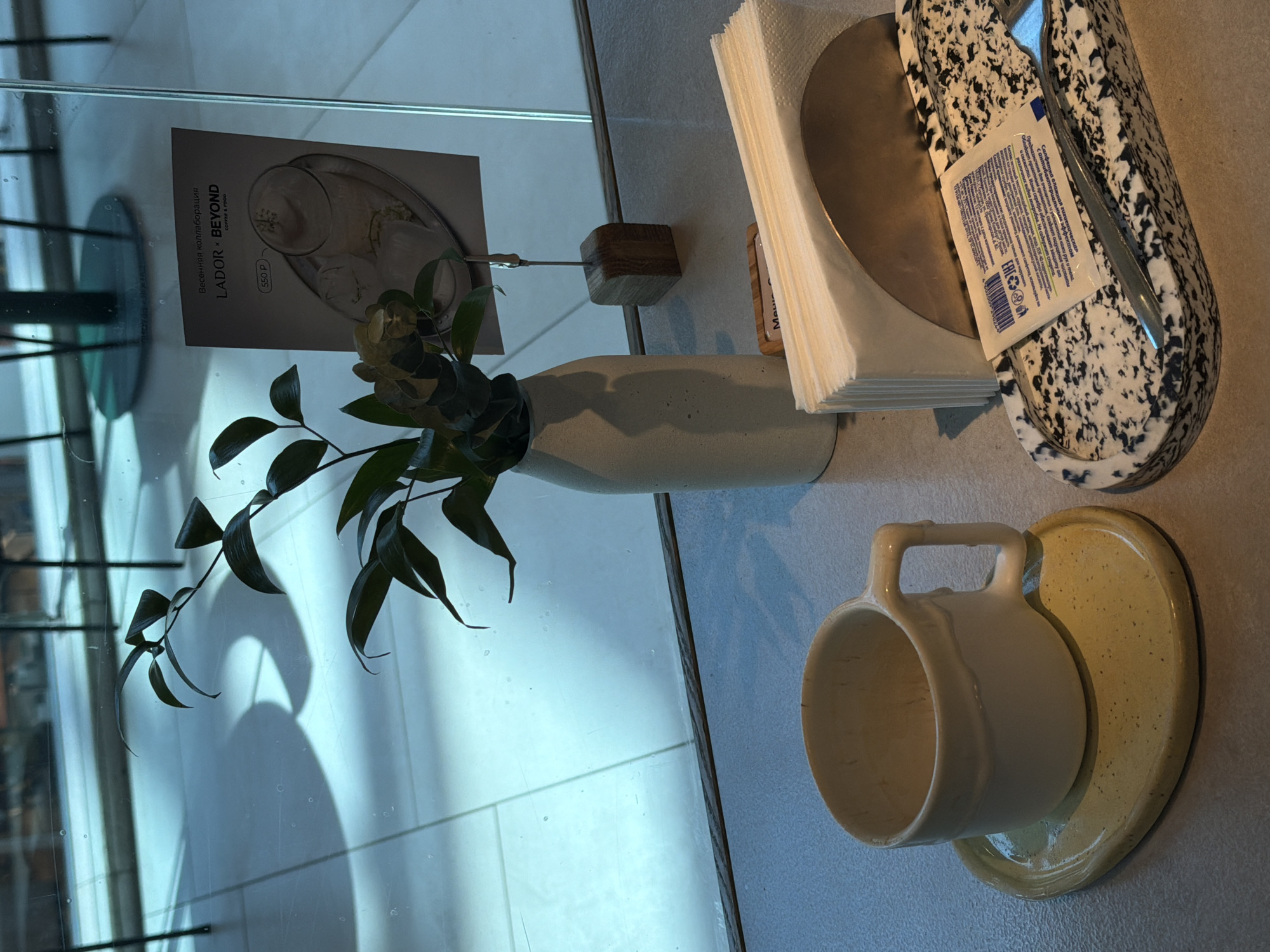 The media file captures an indoor scene, likely a cafe or restaurant table in Khimki, Russia, viewed from a slightly elevated angle. The table is positioned next to a large glass partition, revealing reflections of the interior and casting shadows from objects like the plant.
On the table, a minimalist white ceramic vase holds several dark green leafy branches. To its right, a small wooden block with a metal clip displays a printed card. The card features Russian text reading "Весенняя коллаборация" (Spring collaboration), "LADOR BEYOND", and "COFFEE & FOOD", along with a price of "550 P" (Rubles) next to an image of a drink.
Further right, a stack of white paper napkins rests on a circular metal base. Adjacent to the napkins is a distinctive speckled black and white holder containing cutlery, with a wet wipe packet placed on top. The packet's Russian text indicates it is a "влажная гигиеническая салфетка" (moist hygienic wipe) with an "антибактериальным эффектом" (antibacterial effect), designed for "увлажнения кожи рук и тела" (moisturizing hand and body skin), "без спиртос" (alcohol-free), and contains "Ионы серебра" (silver ions). An "EAC" mark is also visible.
In the foreground, a creamy white ceramic mug with a handle sits on a textured, speckled yellow saucer. The scene is illuminated by natural light, suggesting it is daytime, and casting soft shadows that add depth to the composition. No people are visible in the image.