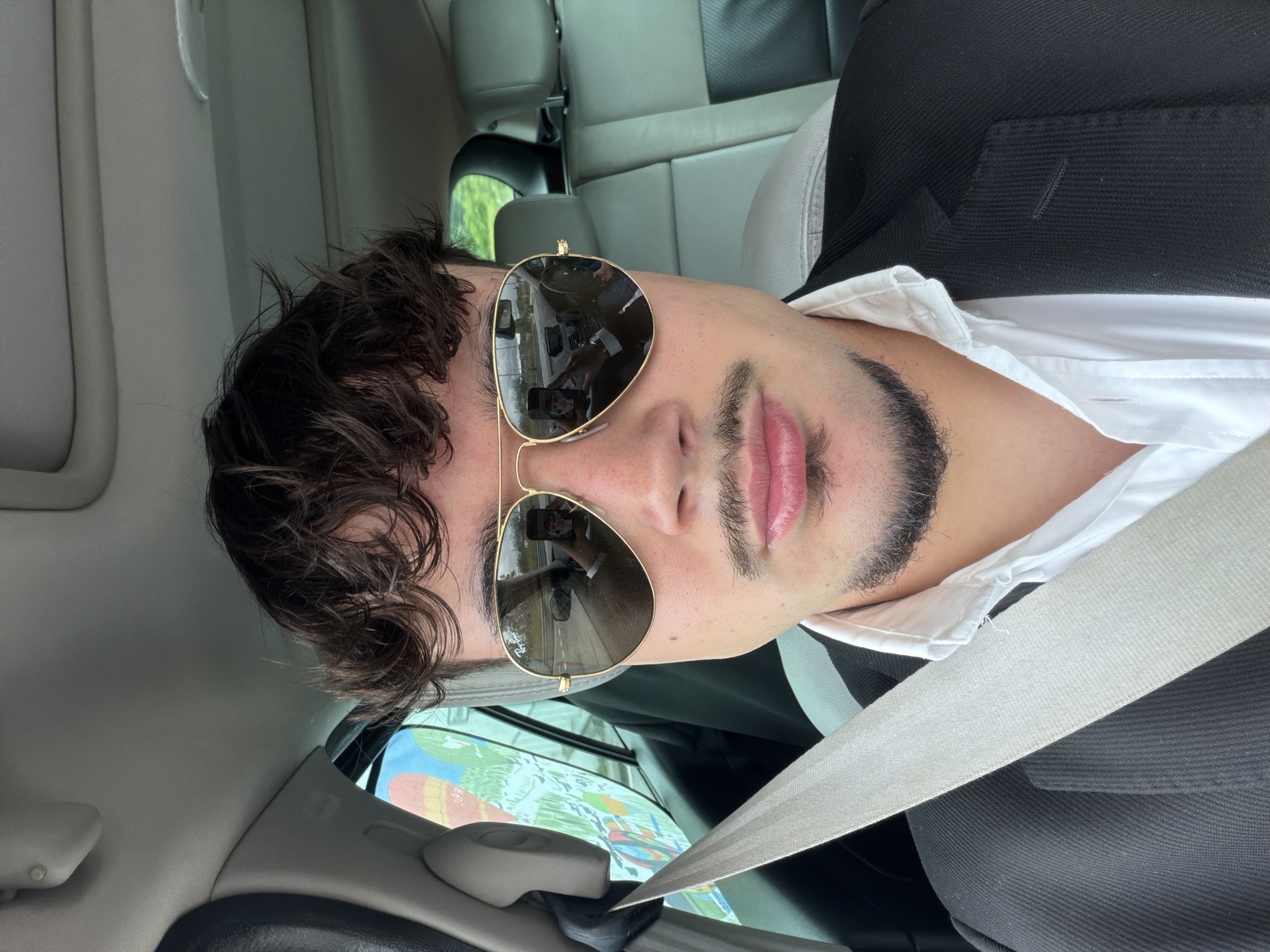 The image depicts a man in a car, wearing a white shirt, black jacket, and gold-rimmed sunglasses. He is also wearing a seatbelt. The reflection in his sunglasses shows a highway scene. The background is blurred, but the interior of the car is visible, including gray leather seats. The location is implied to be near Vila Real, Portugal, based on the highway visible in the sunglasses.