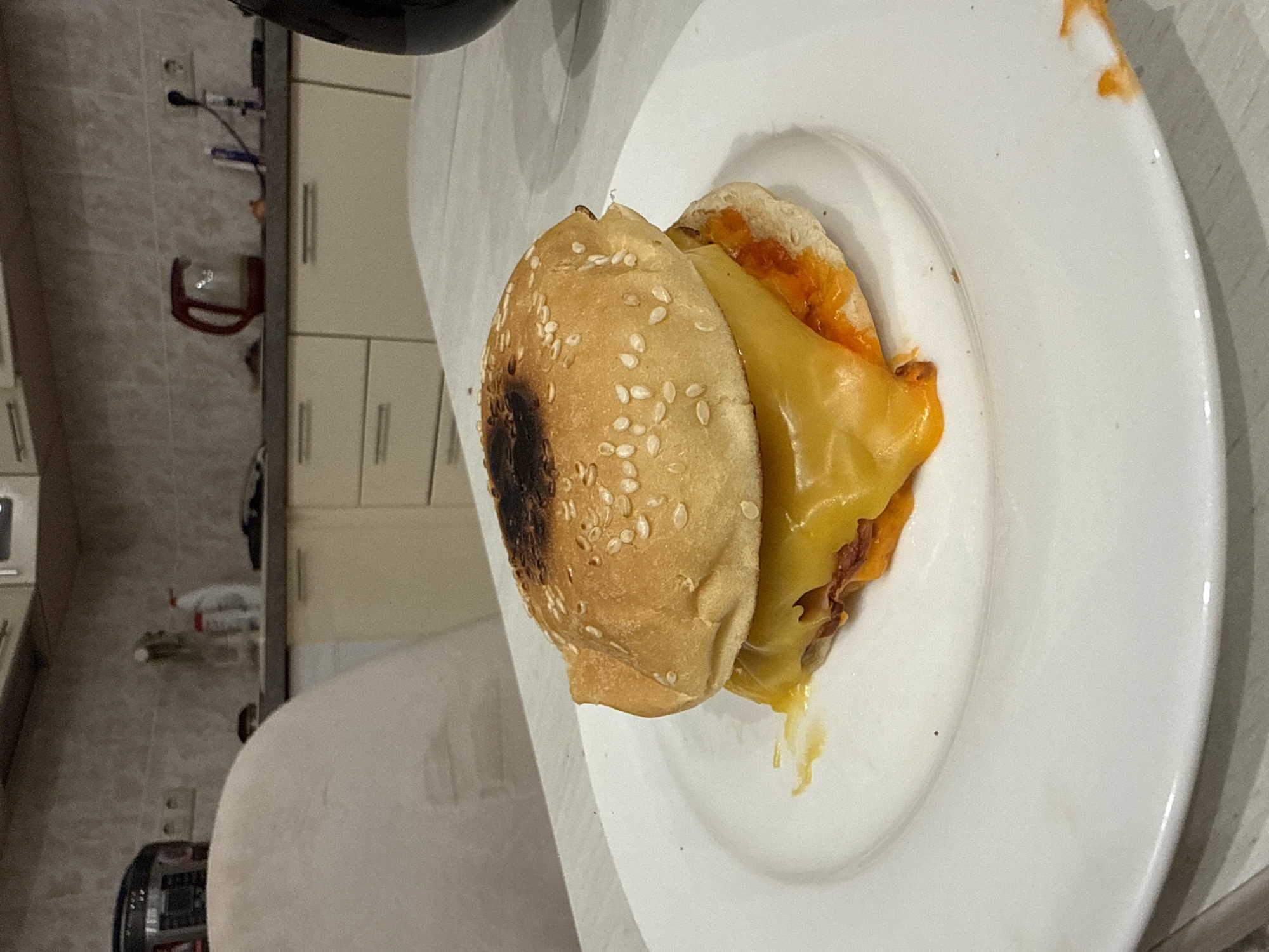 A sesame seed bun sandwich with melted cheese on a white plate is on a surface in a kitchen. The top bun is slightly burned. The location is unspecified, and there's no evidence of it being in Перепечино, Russia.