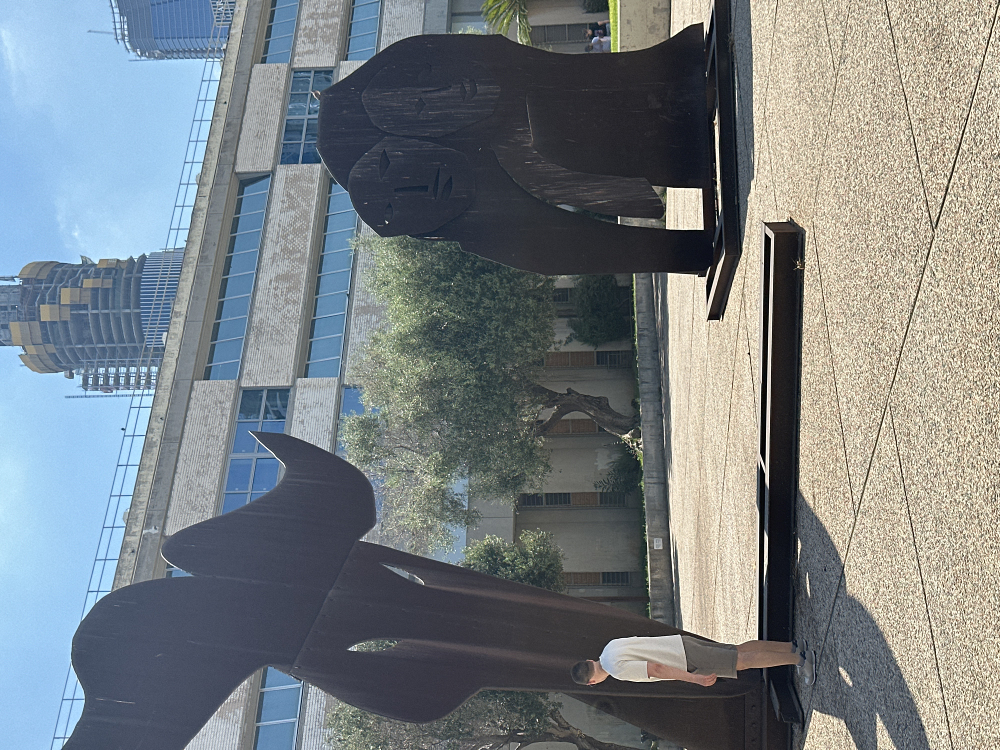 The image depicts an outdoor plaza with two large, dark metal sculptures. In the foreground on the left, a young man wearing a white t-shirt and grey shorts stands facing a sculpture. The sculpture on the left is abstract and appears to be made of curved metal plates. The sculpture on the right is anthropomorphic, resembling two faces joined together. A building with large windows and light-colored brickwork forms the background. In the far background, construction cranes are visible above other buildings, suggesting a city environment. The ground is paved with light-colored tiles. The scene is lit by bright sunlight, casting shadows. This plaza is located at the Tel Aviv University campus.