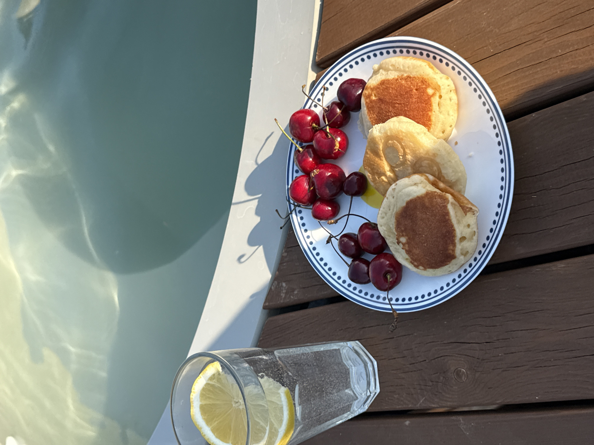 A plate of four mini pancakes with a serving of cherries rests on a wooden deck next to a glass with lemon slices. The scene is set outdoors, near a body of water, possibly a pool or a lake, with light reflecting on the surface. The lighting suggests it is daytime, likely late afternoon, due to the warm, directional sunlight casting shadows. The overall ambiance is relaxed and leisurely, suggesting a meal or snack enjoyed in a tranquil outdoor setting. The plate has a blue and white patterned rim. The cherries are a deep red color. The glass is clear and filled with a clear liquid, possibly water or a light beverage, with two thin slices of lemon visible. The wooden deck has distinct grain patterns and a warm brown hue. There is no visible text in the image.