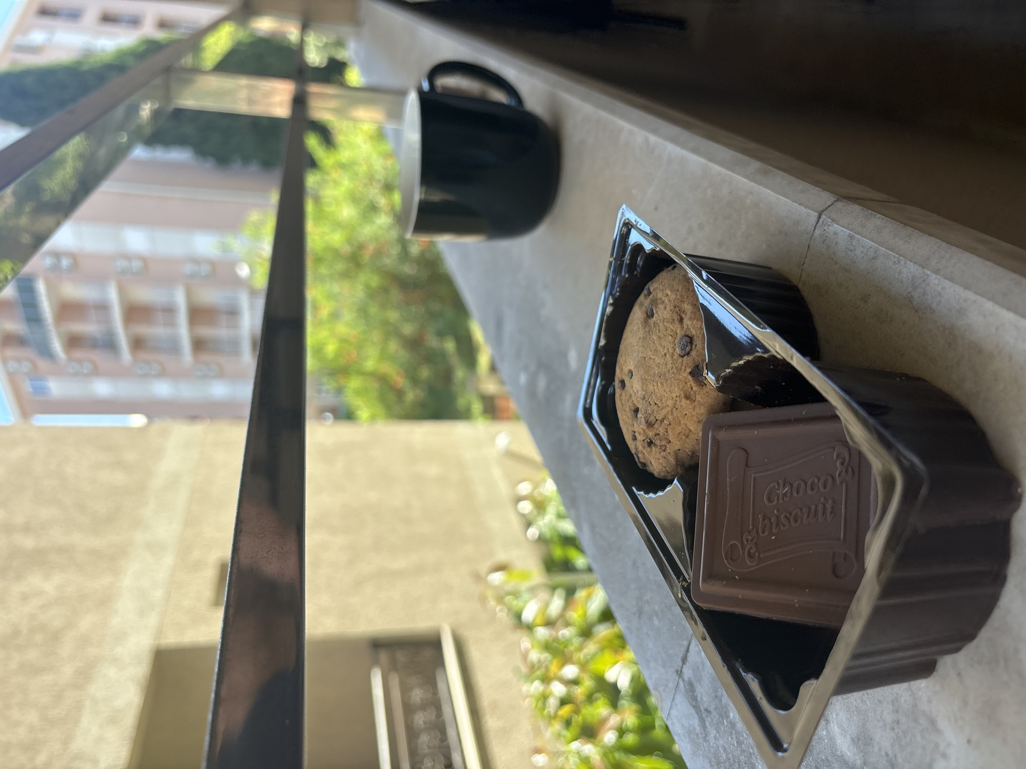 A partially eaten cookie and a Choco Biscuit bar sit in a black plastic container on a ledge. Next to the container is a dark mug. In the background, a building with multiple windows is visible, along with green trees and foliage. The scene is likely located in Budva, Montenegro. A portion of a metal railing is visible on the left side of the image.