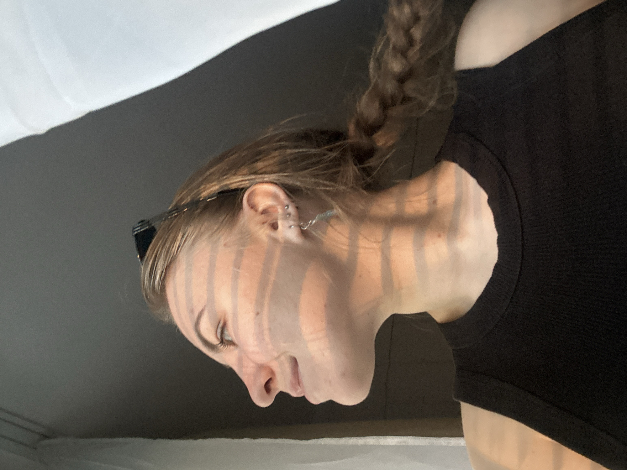 A close-up, vertically oriented image features a person's head, neck, and upper shoulder against a blurry background of white and gray tones. The subject, appearing to be a young woman or non-binary individual, is shown in profile, looking downwards towards the bottom of the frame. Their light brown or blonde hair is partially pulled back in a braid, with some strands framing their face. They are wearing a black ribbed tank top or camisole.
Notable details include multiple ear piercings, specifically a helix piercing and two lobe piercings, one of which has a dangling earring with a chain-like detail. What appears to be sunglasses or a hair clip is pushed back into the hair above the ear. Distinct vertical striped shadows are cast across the person's face, neck, and shoulder, suggesting strong, directional light coming through an object like window blinds. The light source implies a daytime setting, likely indoors. The expression is neutral, with a soft downward gaze. There is no visible text in the image. The setting is an unknown indoor location.