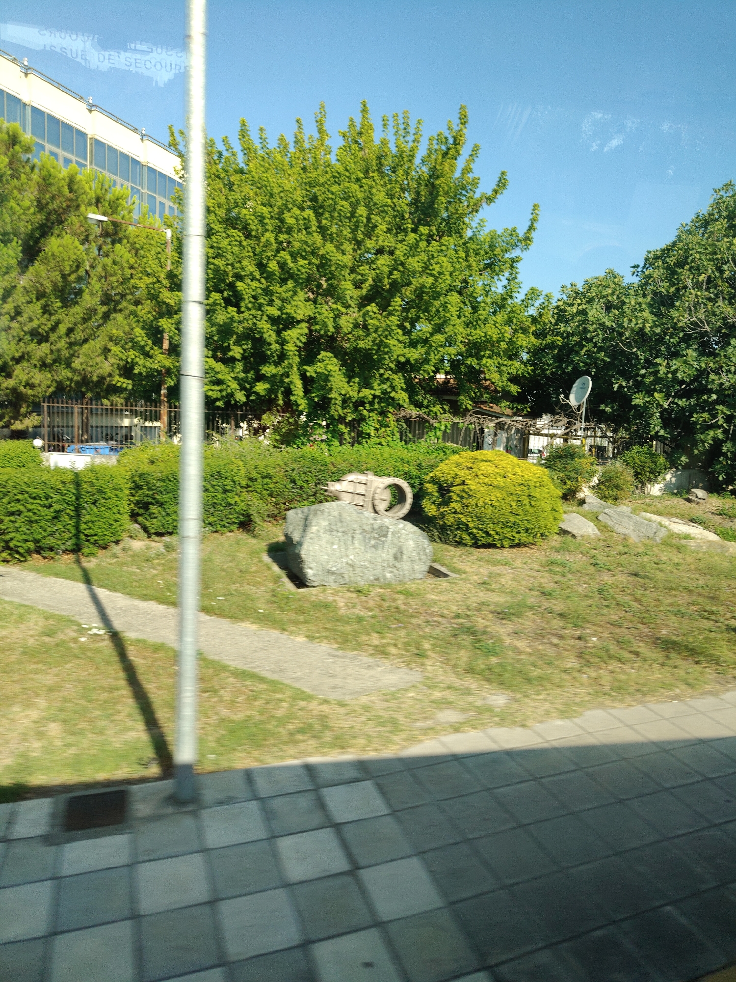 The image captures a sunlit outdoor scene, likely in Larissa, Greece, showcasing a blend of urban and natural elements. In the background, a modern building with a light-colored facade and numerous windows stands tall. Lush green trees, including a large, dense one, dominate the mid-ground. To the right, a satellite dish is visible, mounted on a pole. In the foreground, a grey, textured paved area transitions to a grassy area with a pathway. A large, grey boulder with a circular metallic object resting on it is a prominent feature in the middle ground. A tall, metallic pole stands vertically in the center of the image, casting a long shadow. The sky is a clear, bright blue. The overall impression is a clean and orderly environment, possibly a public space or park adjacent to a building.